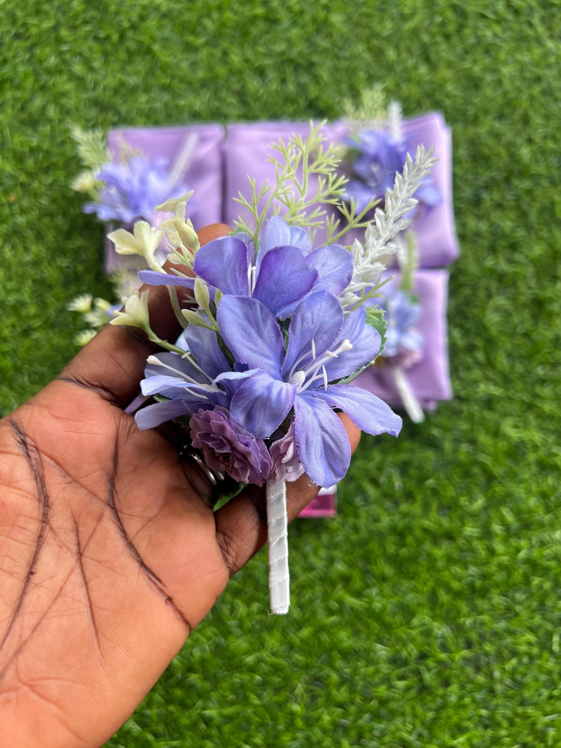 Hand holding a purple floral boutonniere with green sprigs over a green grass background.