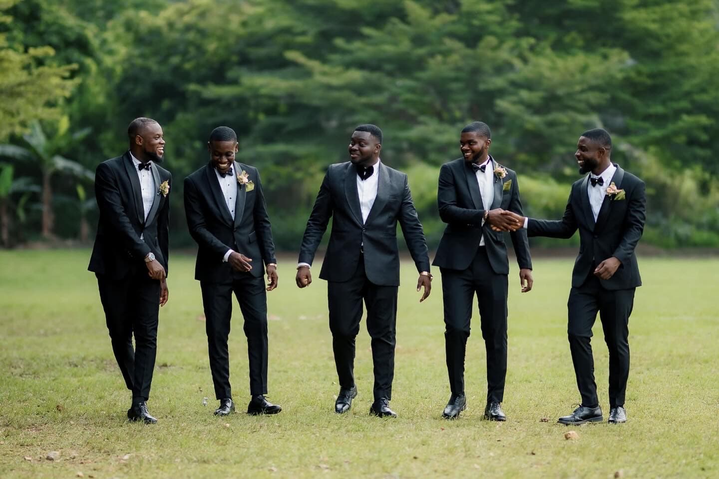 Five men in black tuxedos walk across a green field, smiling and shaking hands warmly.