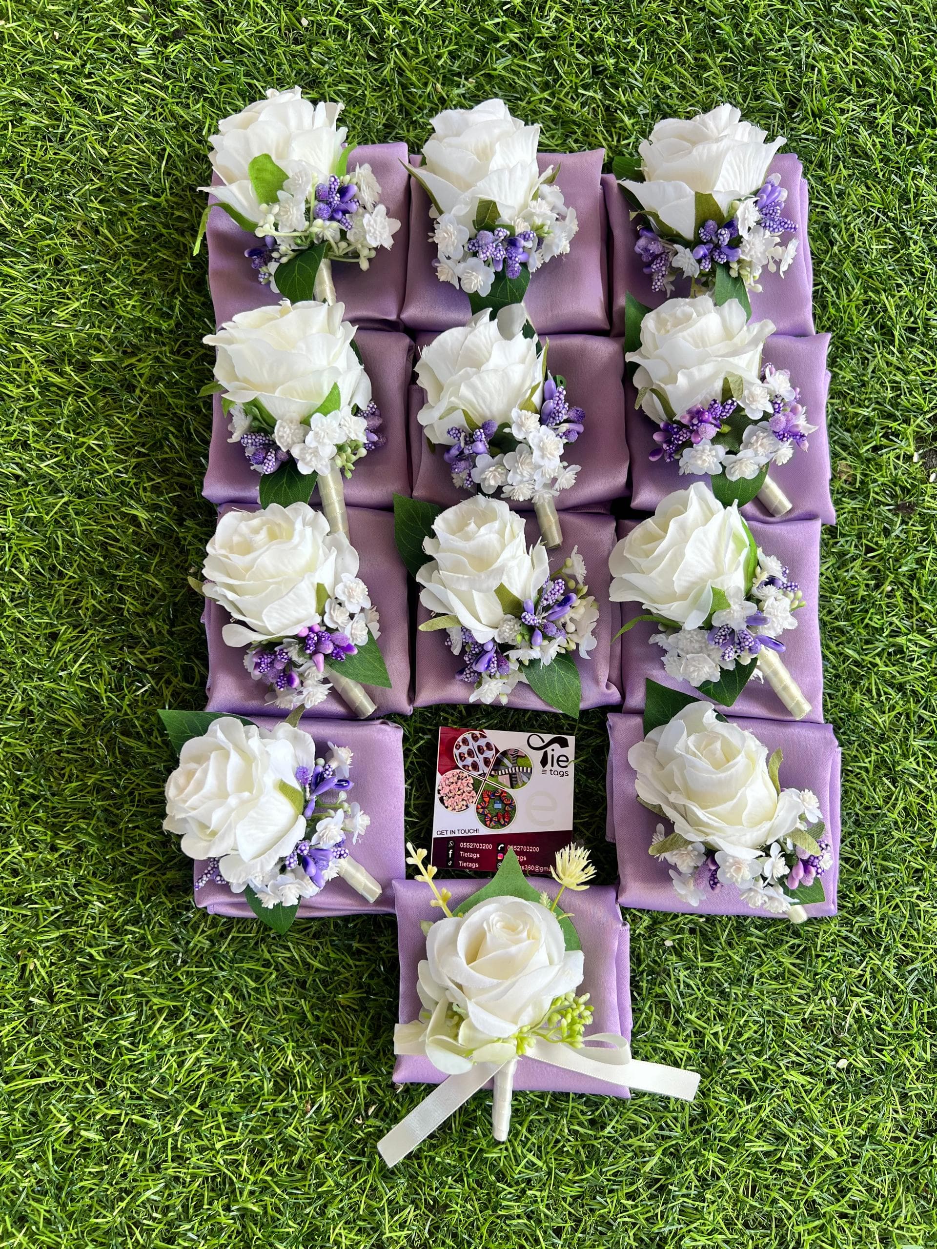 White rose boutonnieres with purple accents on lavender pocket squares arranged on green grass.