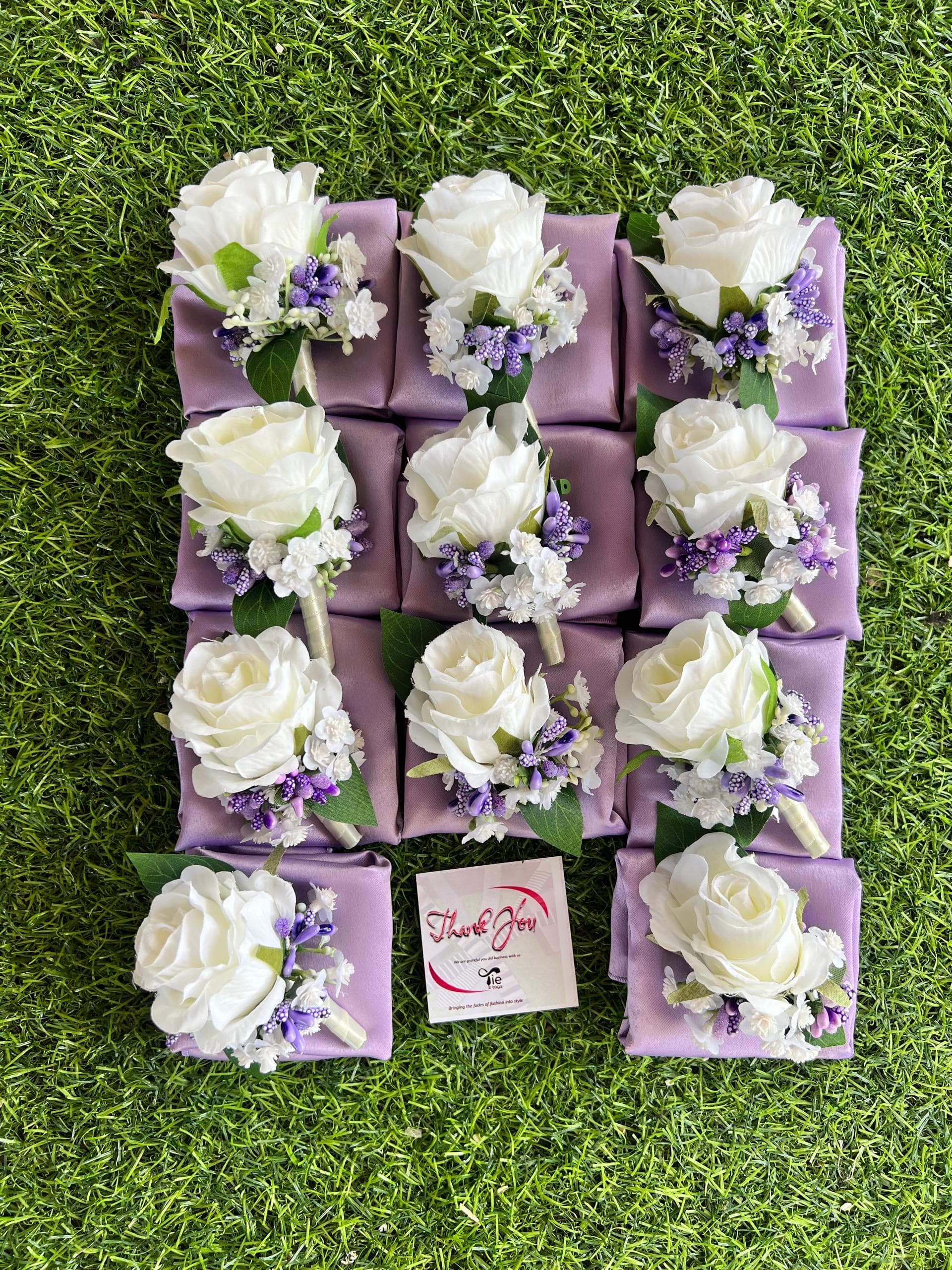 Eleven white rose boutonnieres with purple accents on lavender pocket squares over green grass.