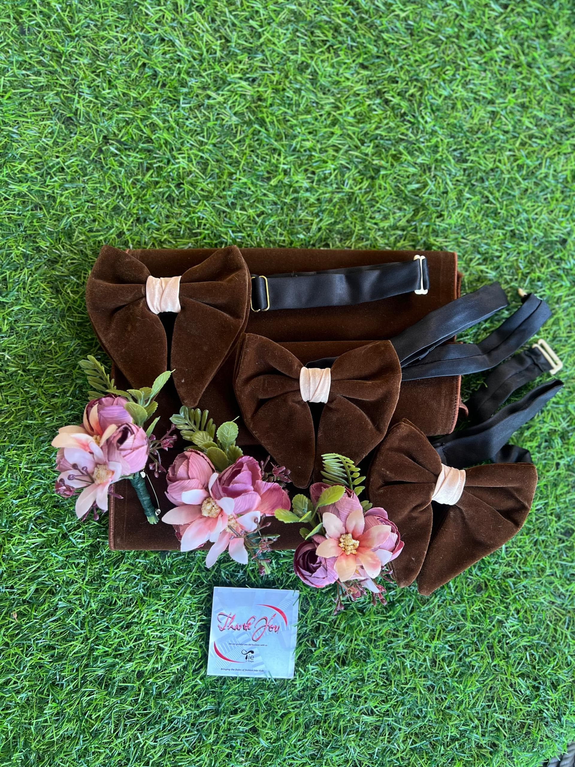Three brown velvet bow ties and pink floral boutonnieres on a box over artificial grass.