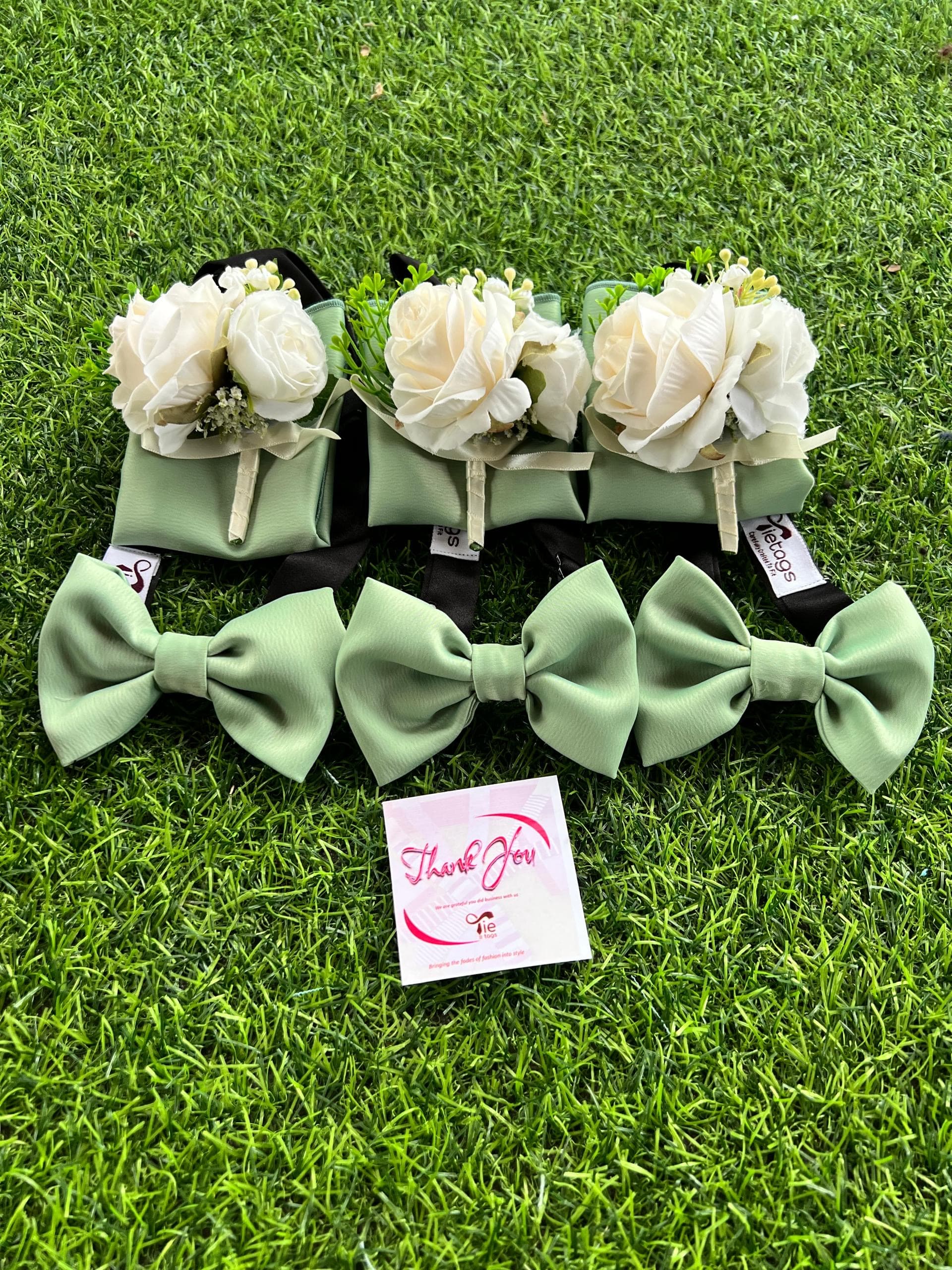 Three sets of sage green bow ties and white rose boutonnieres on green grass.