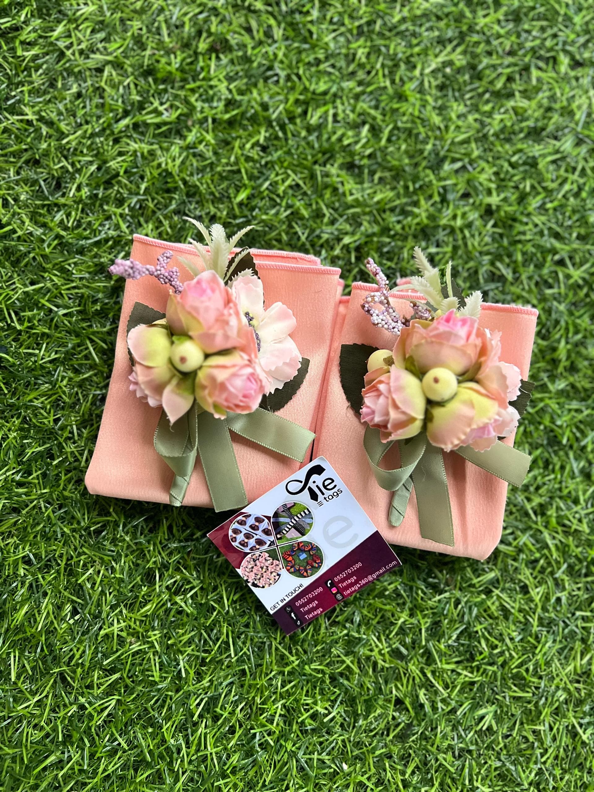 Two peach fabric pouches decorated with pink artificial roses and green ribbons on green grass.