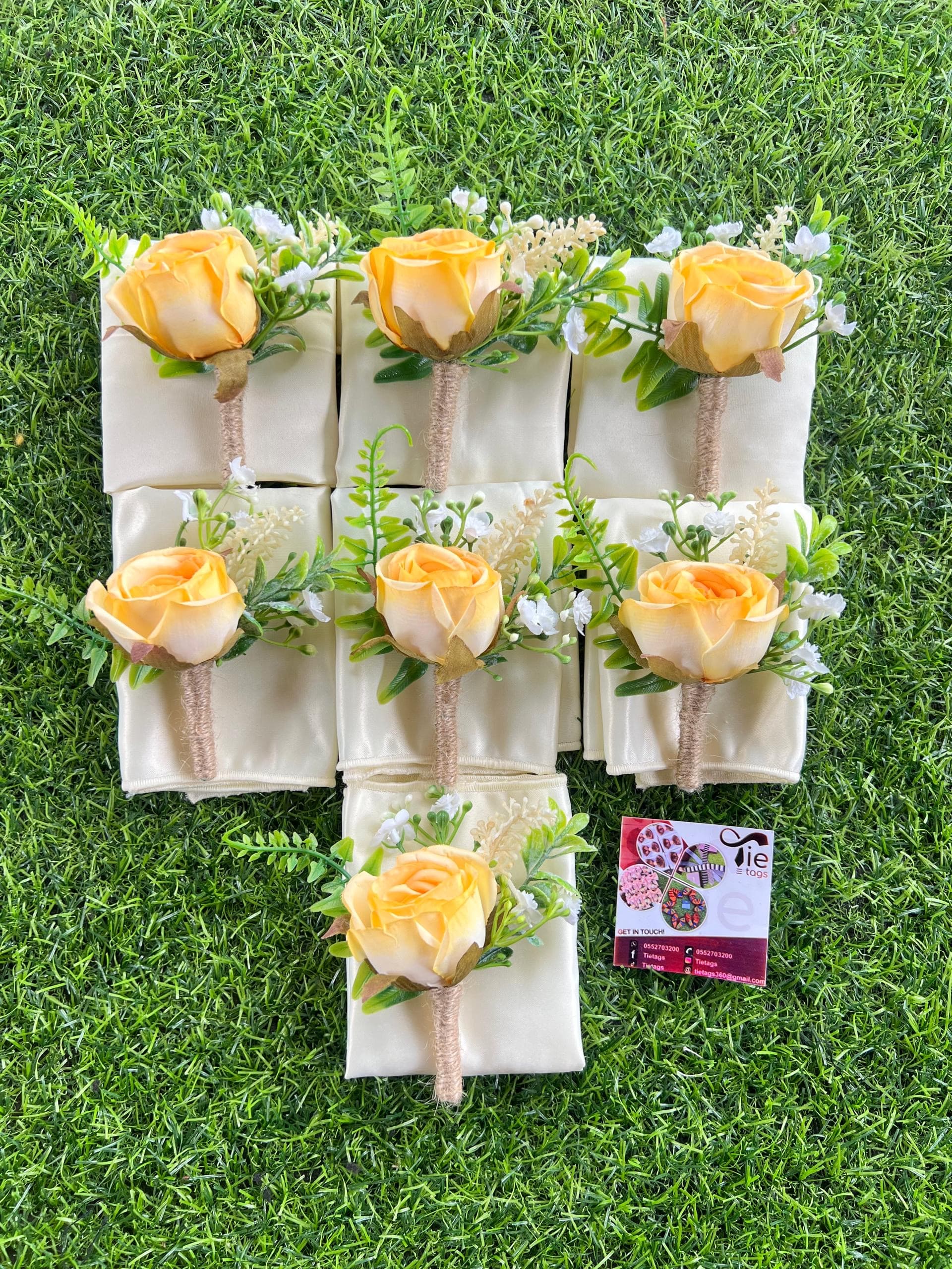 Seven yellow rose boutonnieres with twine-wrapped stems resting on cream fabric over green grass.