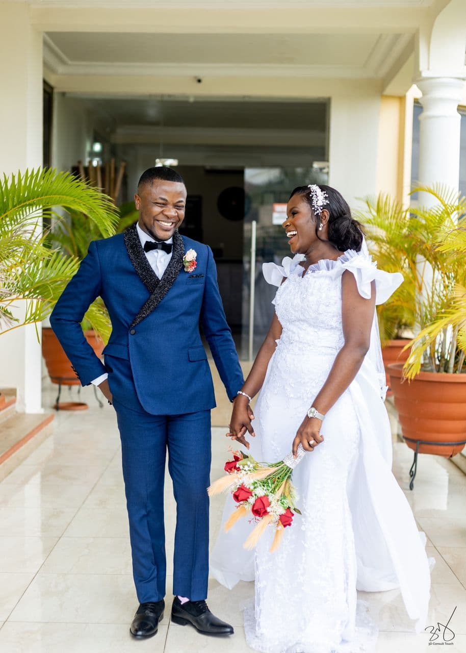 Joyful bride and groom laughing together, holding hands and a bouquet of red roses outdoors.