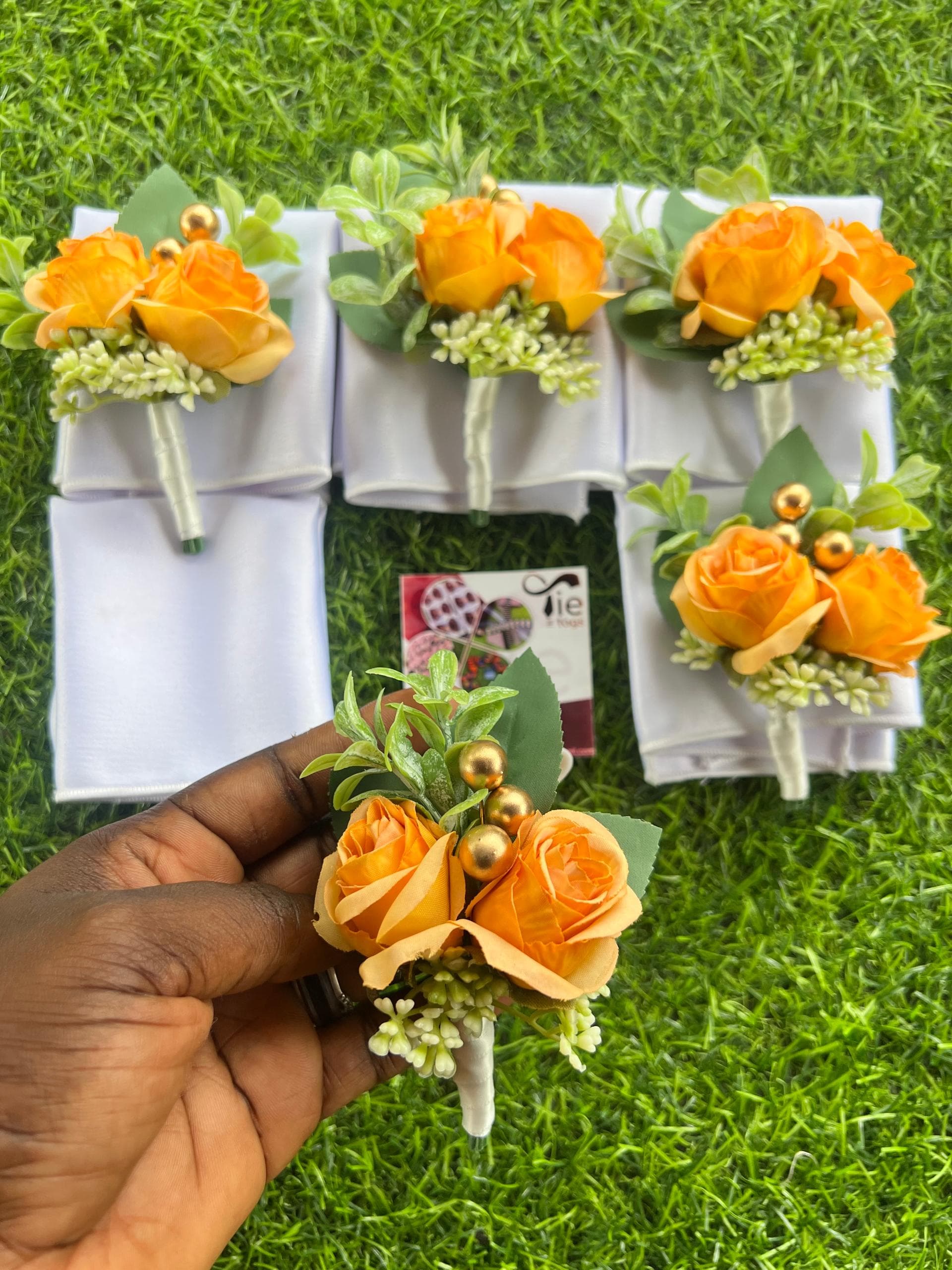 Hand holding an orange rose boutonniere with gold beads over white pocket squares on grass.