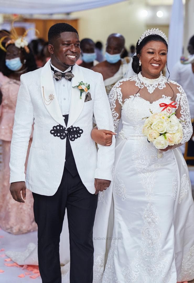 Smiling bride in lace gown and groom in white tuxedo walk down the wedding aisle.