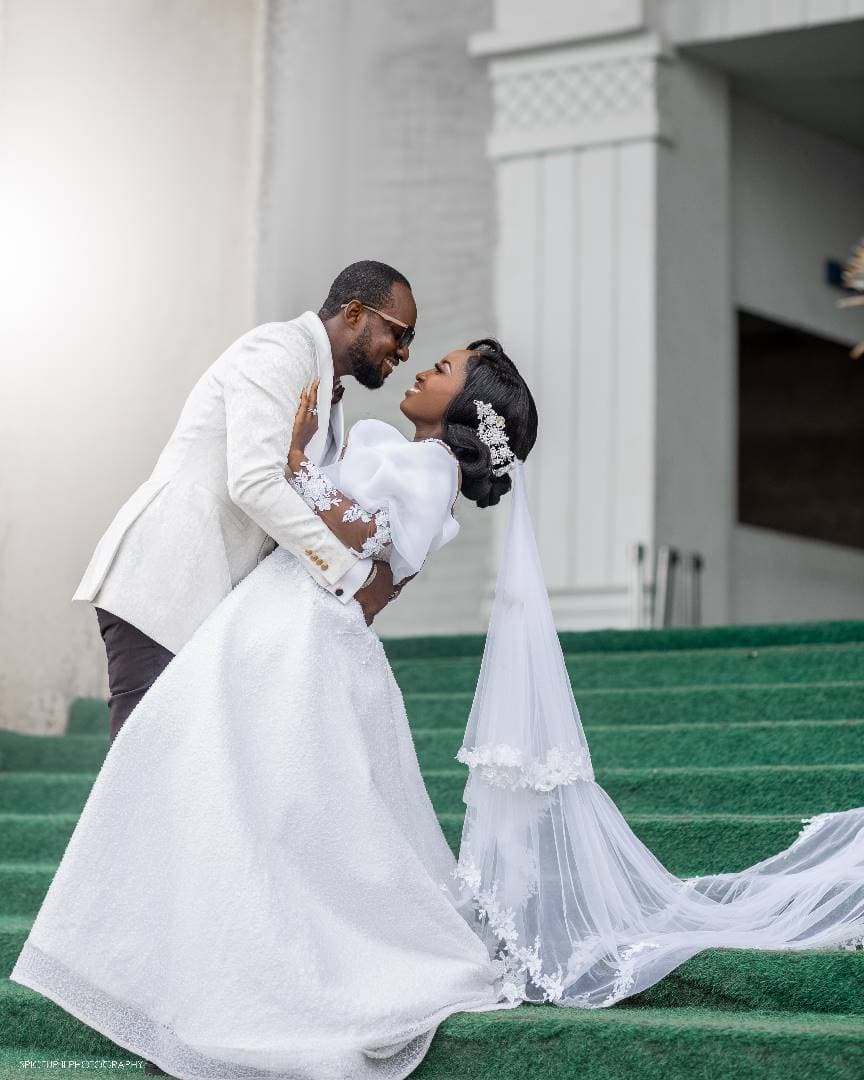 Groom in white suit leans over bride in white gown on green carpeted stairs.