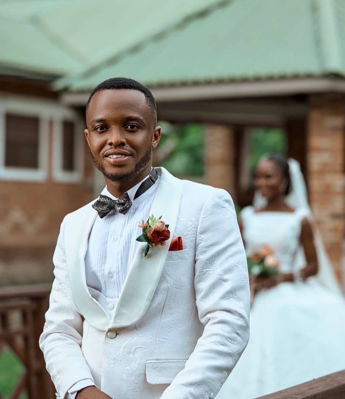 Smiling groom in a white tuxedo with his bride blurred in the background outdoors.