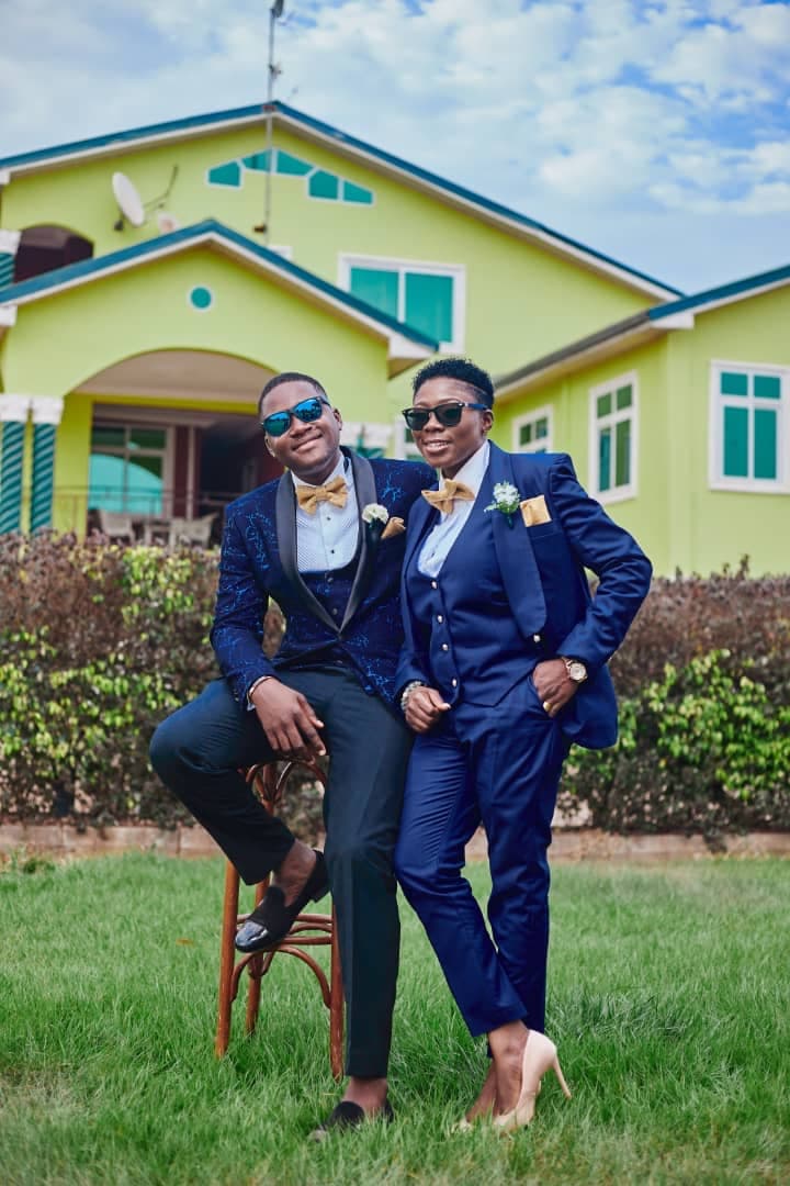 Man and woman in blue suits and gold bow ties pose outside a green house.
