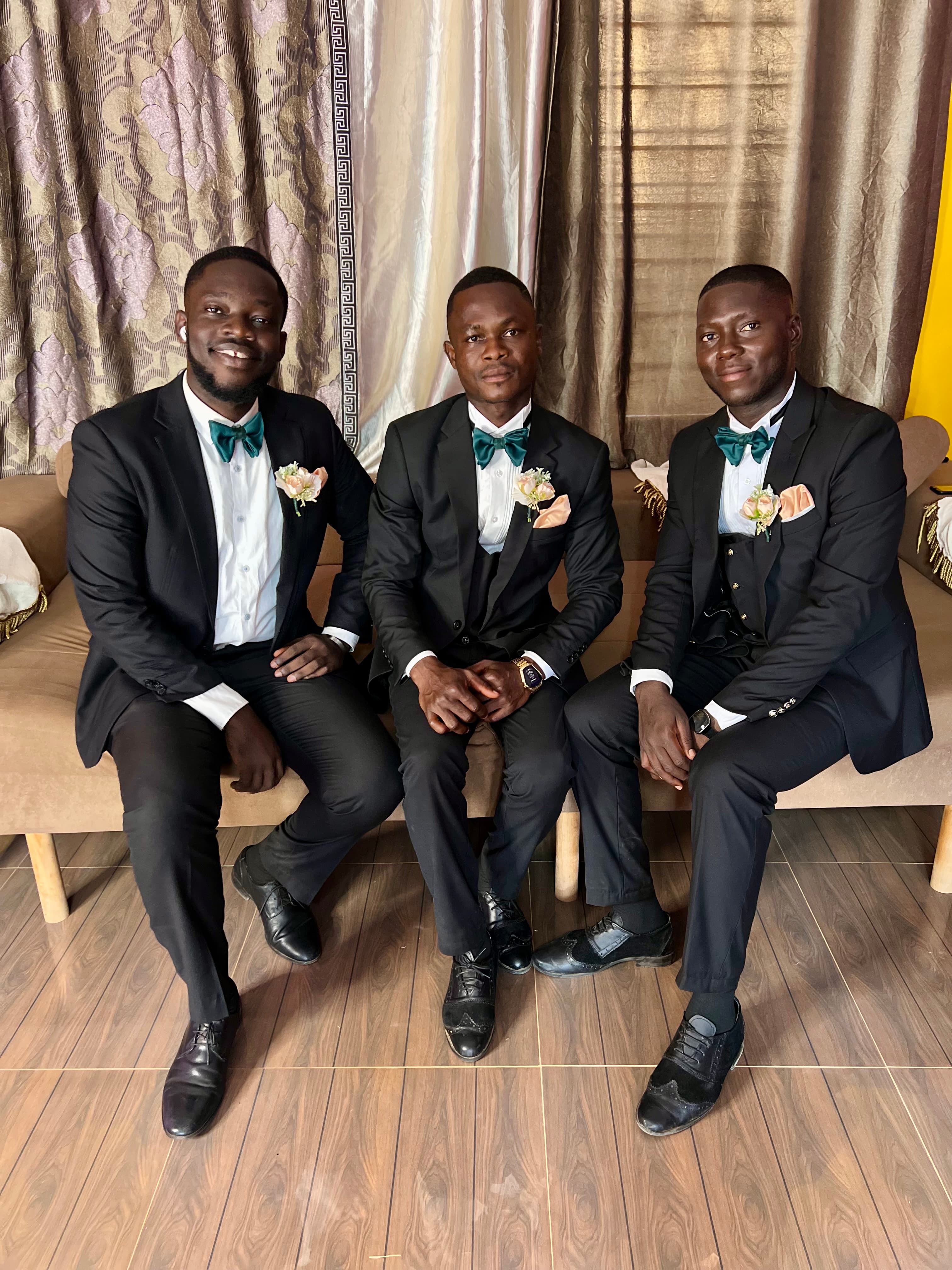 Three men in black suits with green bow ties and boutonnieres sitting on a couch.