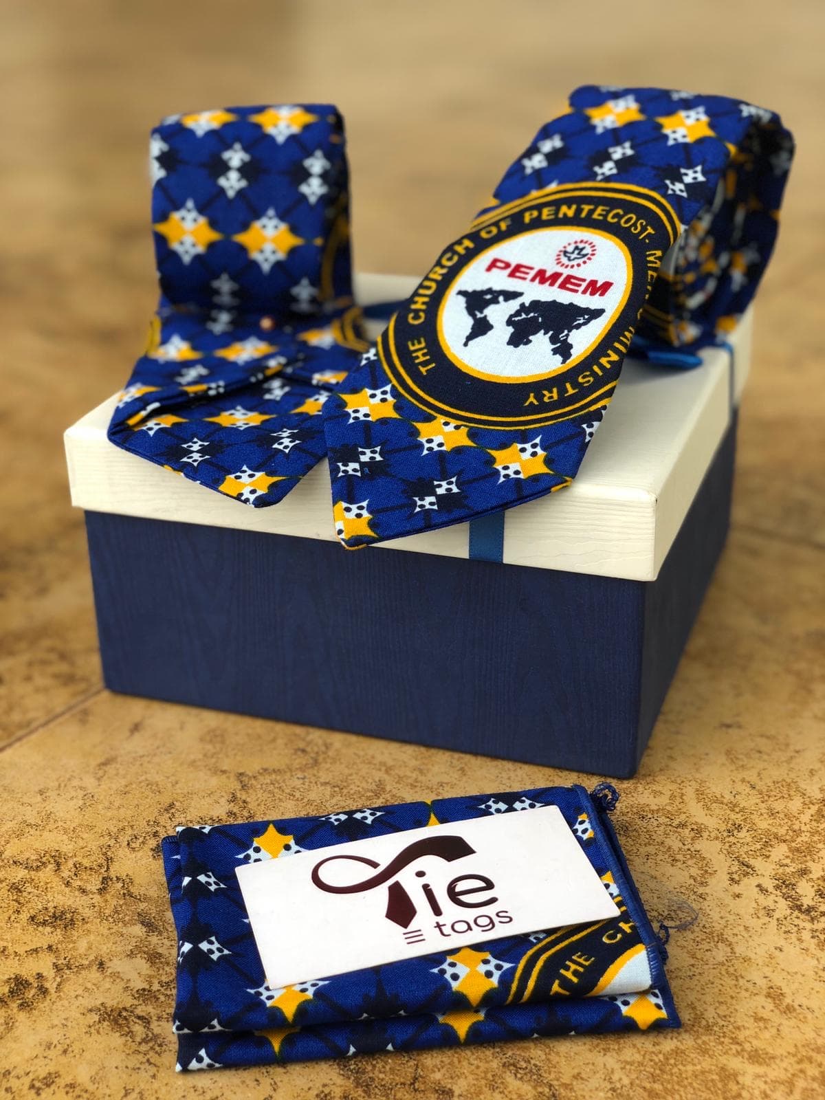 Blue patterned tie with PEMEM logo displayed on a gift box with pocket square.