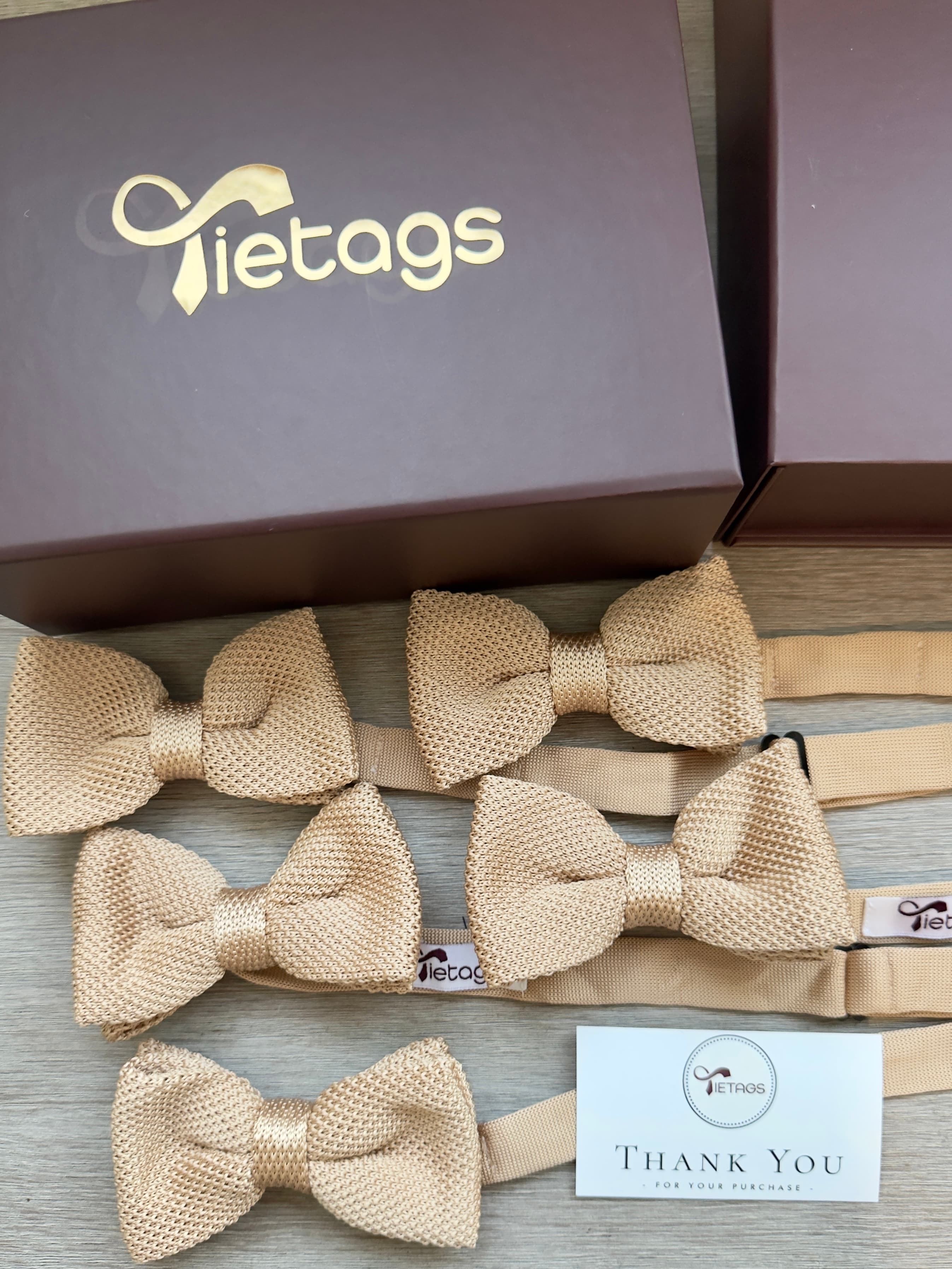Five champagne-colored knitted bow ties arranged beside a brown Tietags box and thank you card.