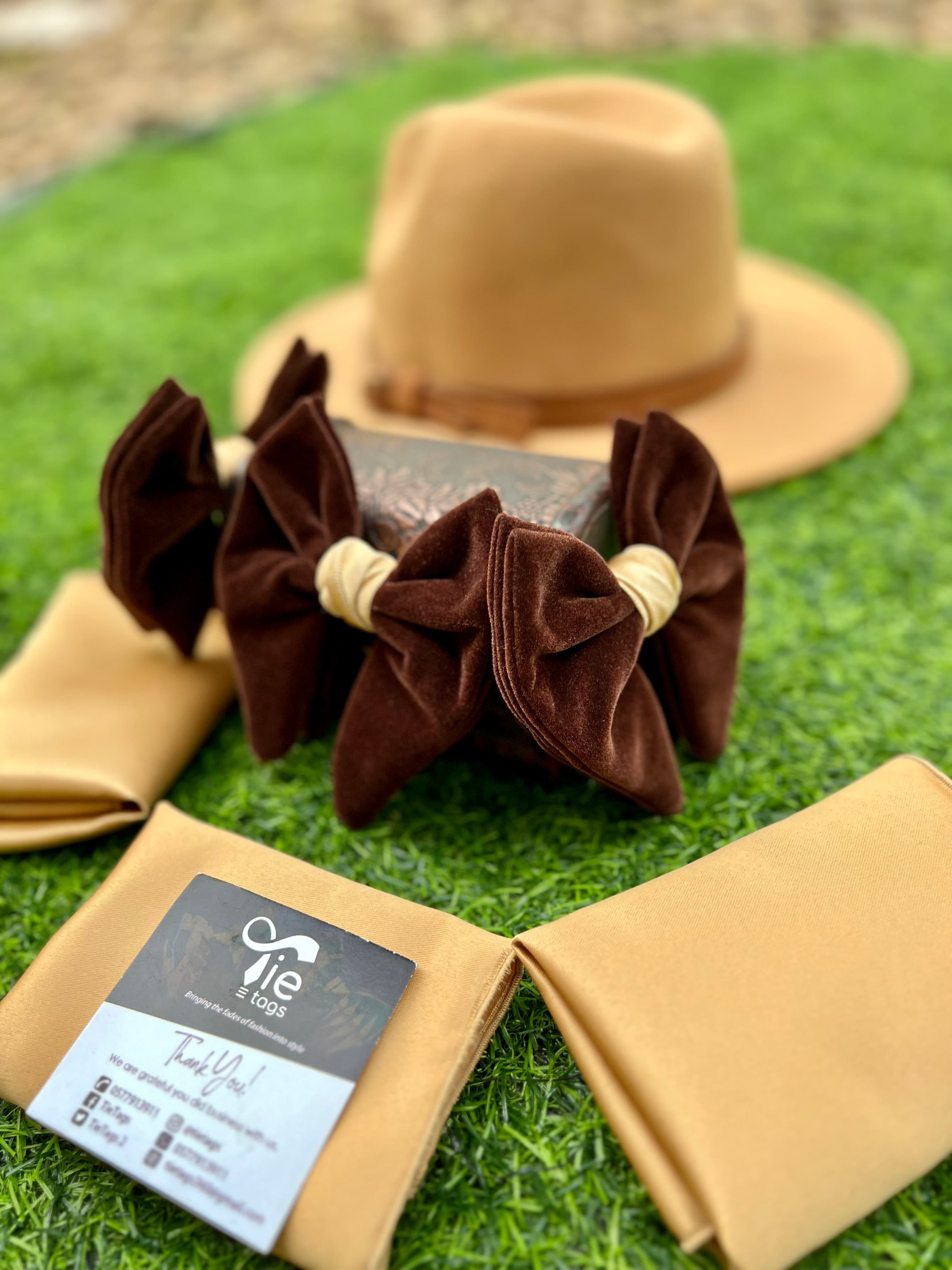 Tan fedora hat, dark brown velvet bow ties, and beige pocket squares on green grass.