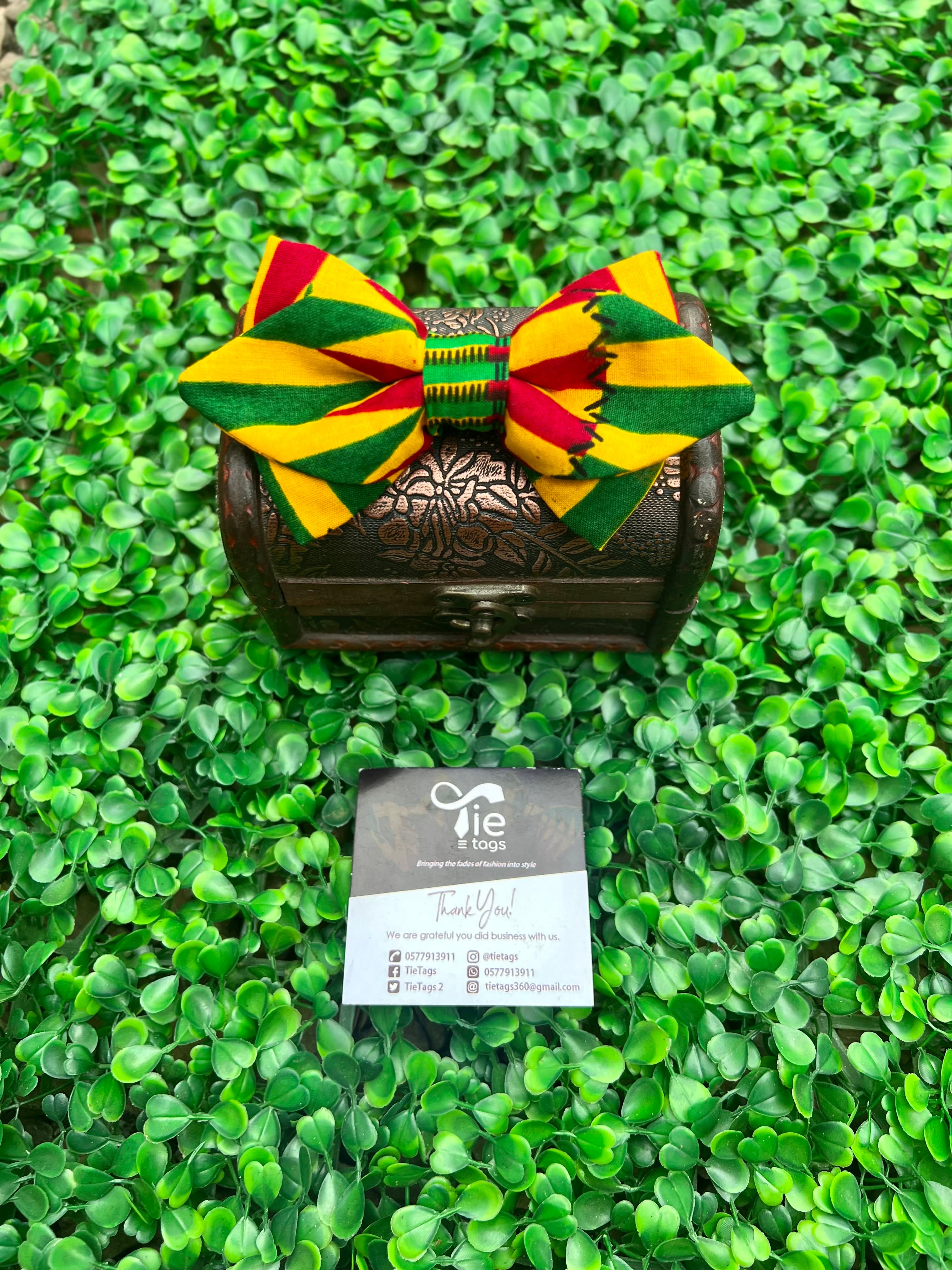 Colorful African print bow tie on a small ornate chest against a green leafy background.