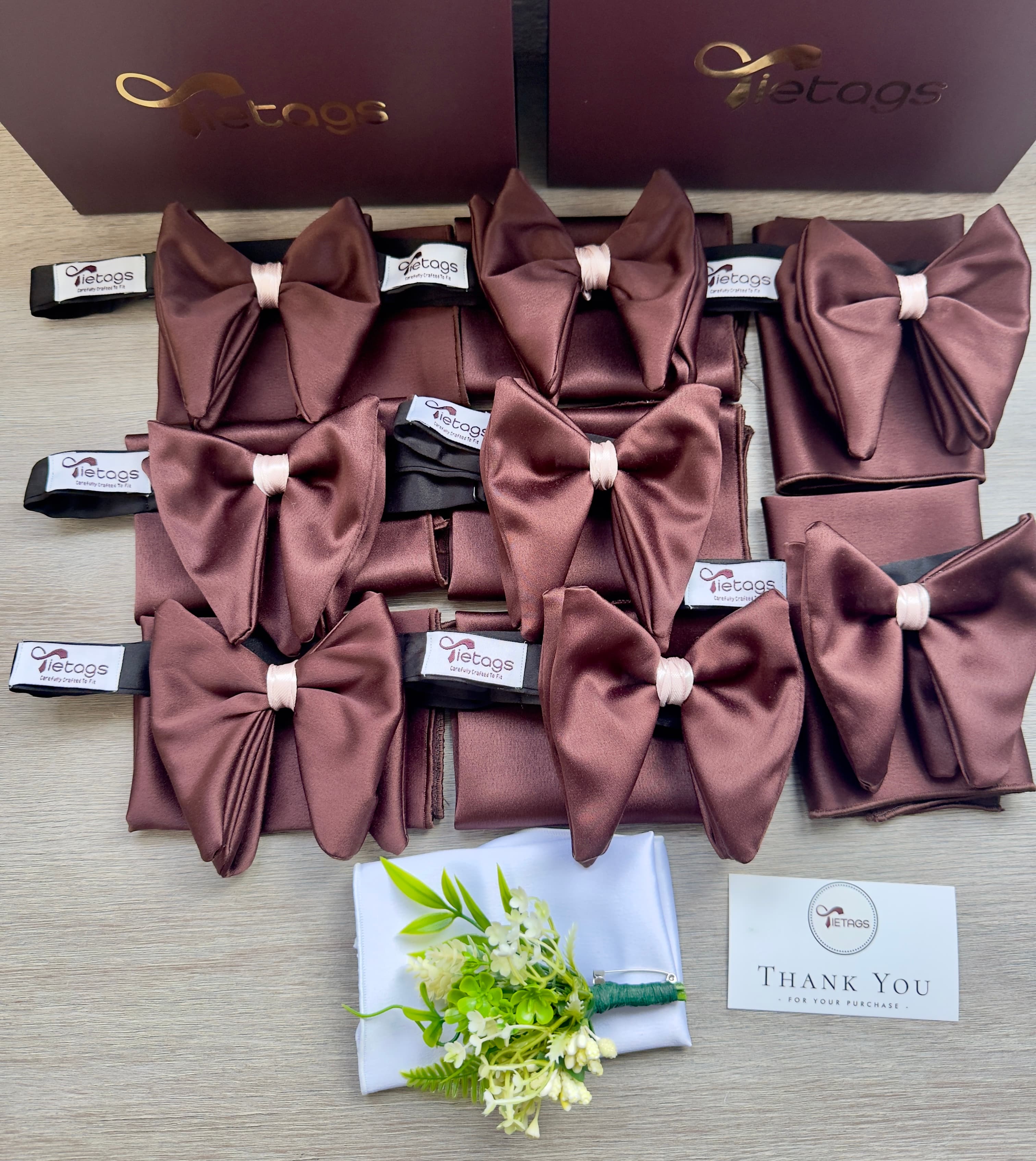 Brown satin bow ties and pocket squares displayed with a floral boutonniere and brand boxes.