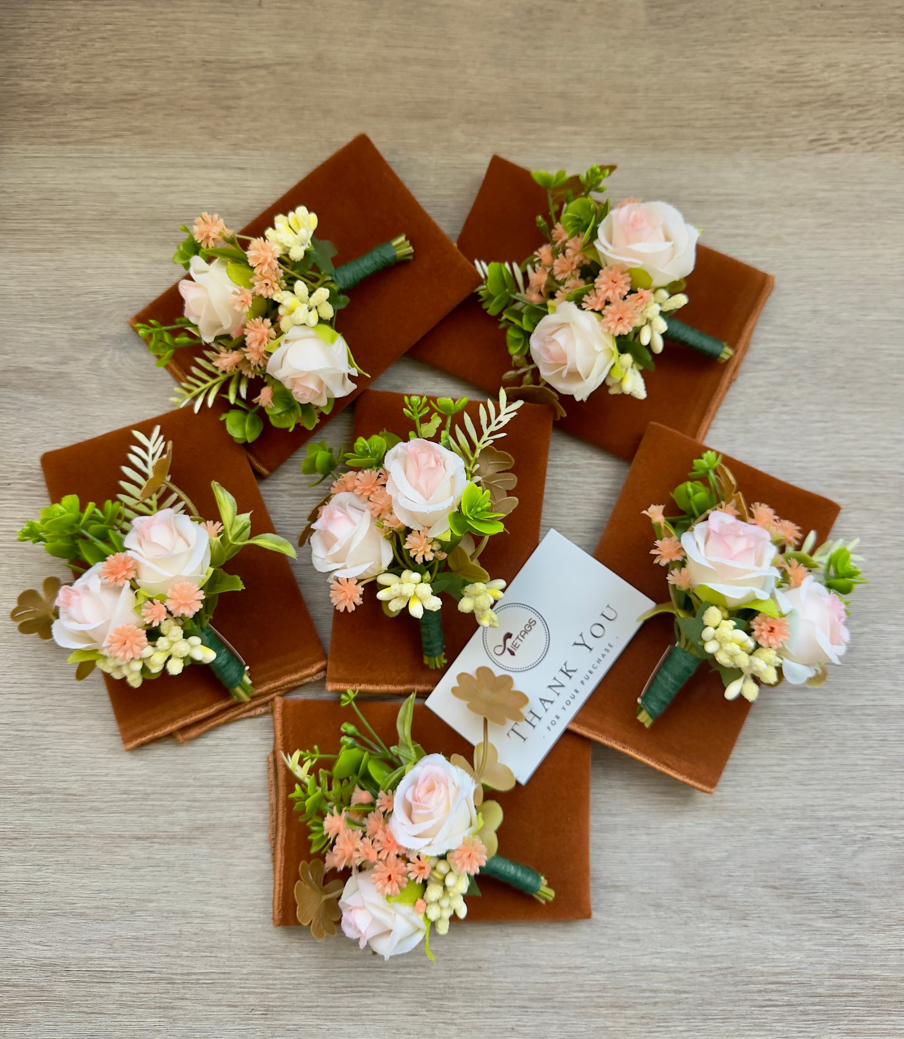 Six pink rose boutonnieres on brown velvet squares arranged around a small thank you card.