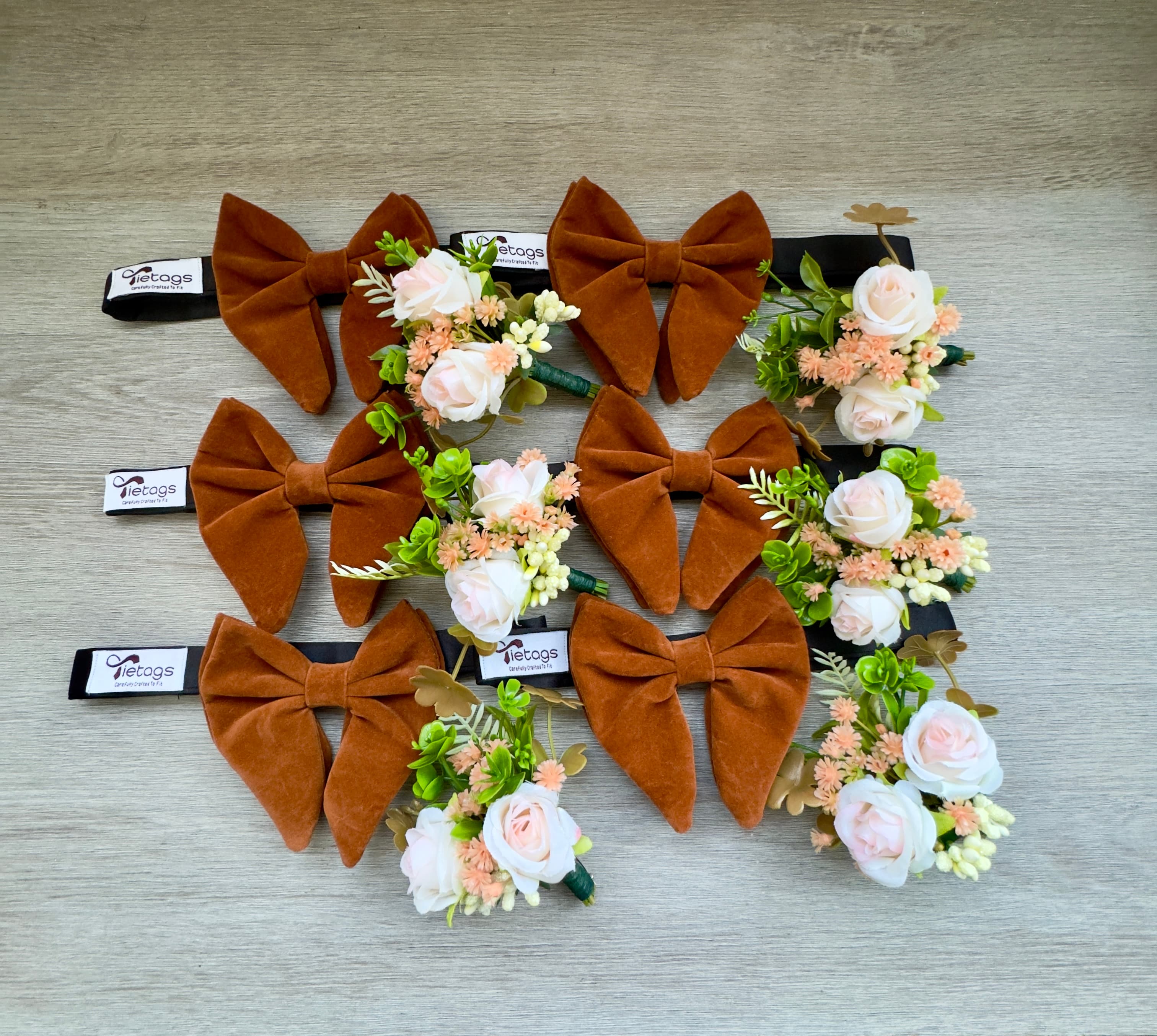 Six rust velvet bow ties and peach and white floral boutonnieres on light wood.