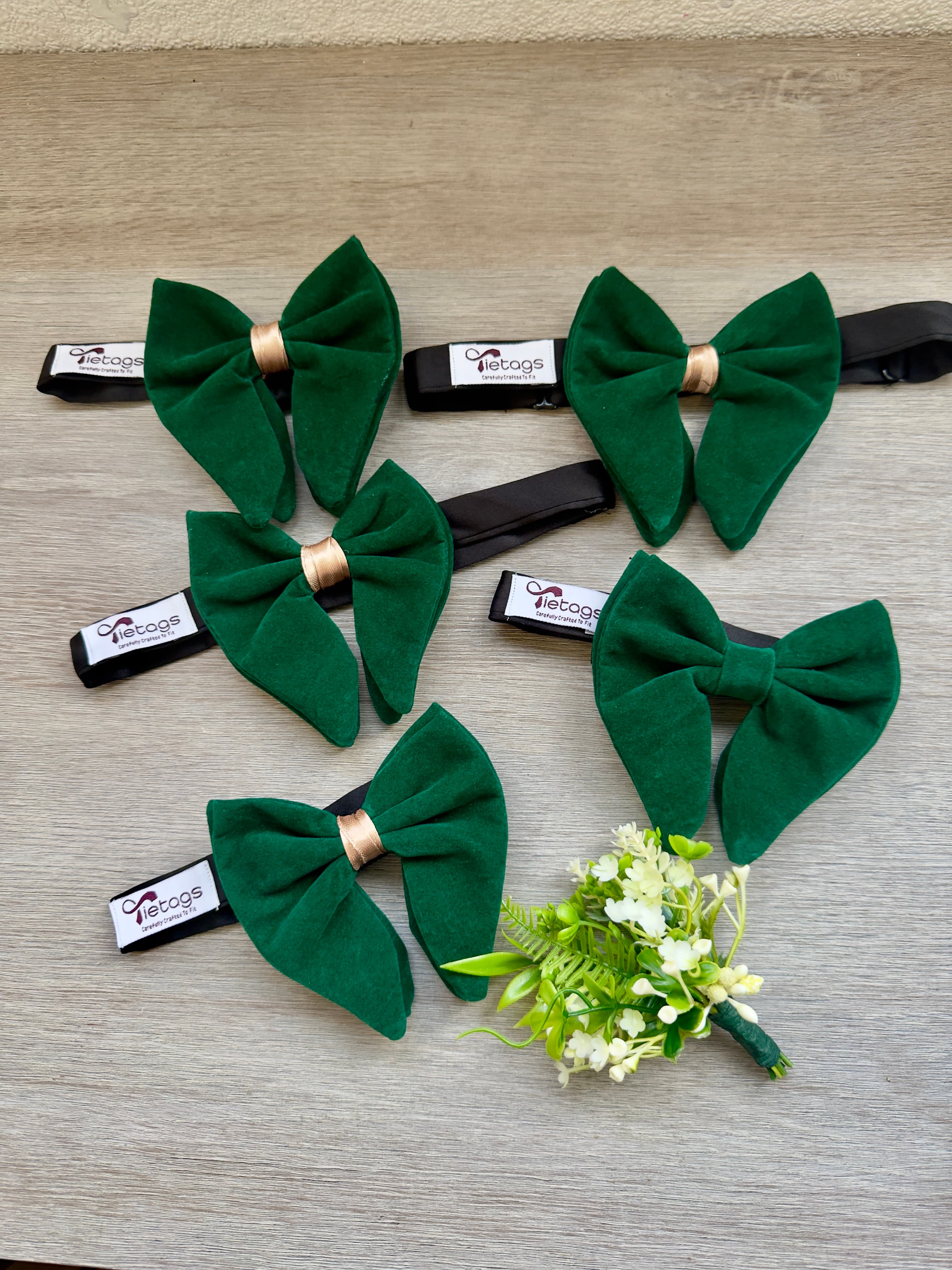 Five green velvet bow ties with gold accents and a white boutonniere on wood.