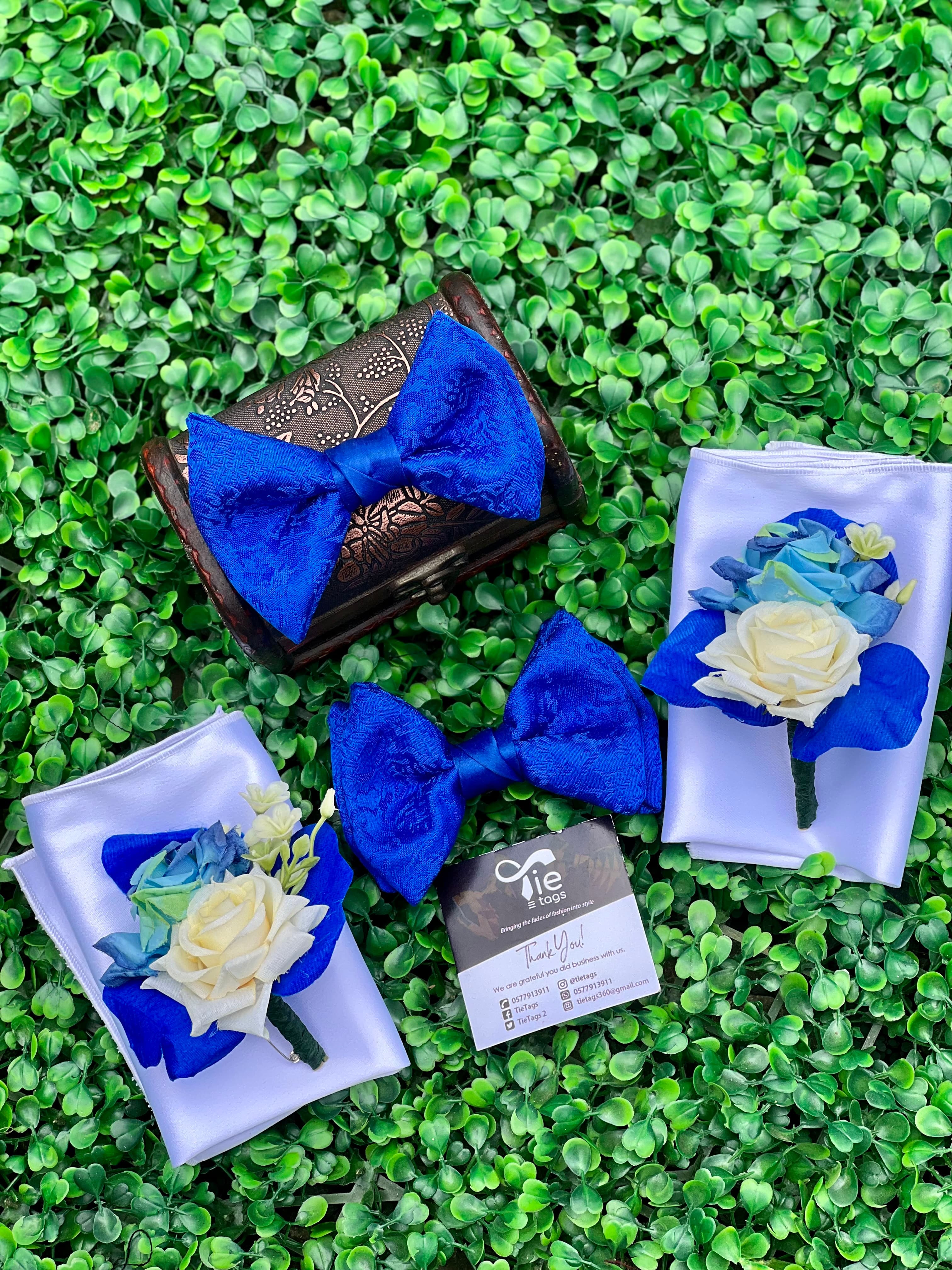 Royal blue bow ties and floral boutonnieres on white pocket squares against green foliage.