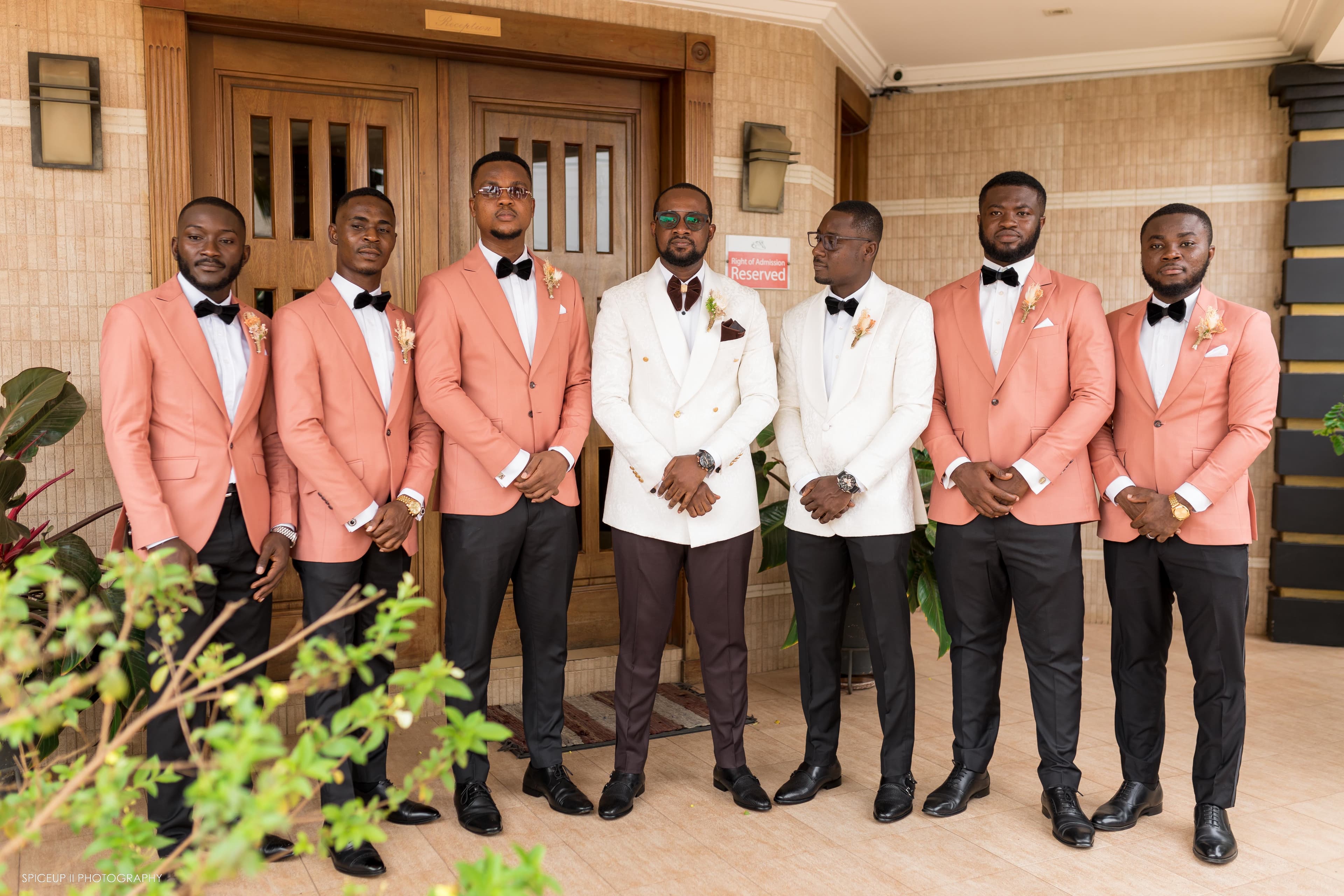 Seven men in peach and white blazers posing outside a wooden reception door.