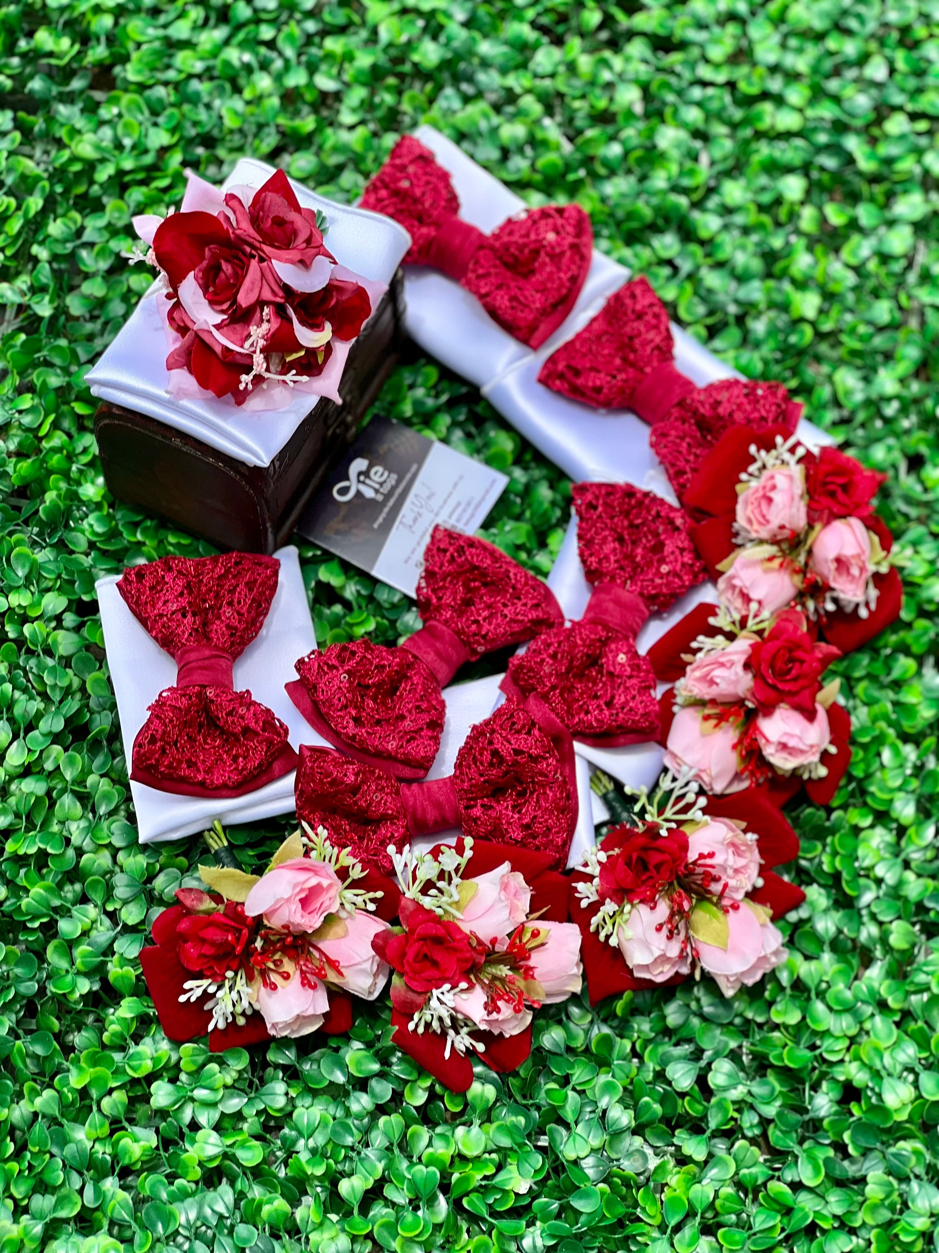 Red lace bow ties and pink floral boutonnieres arranged on a green leafy background.
