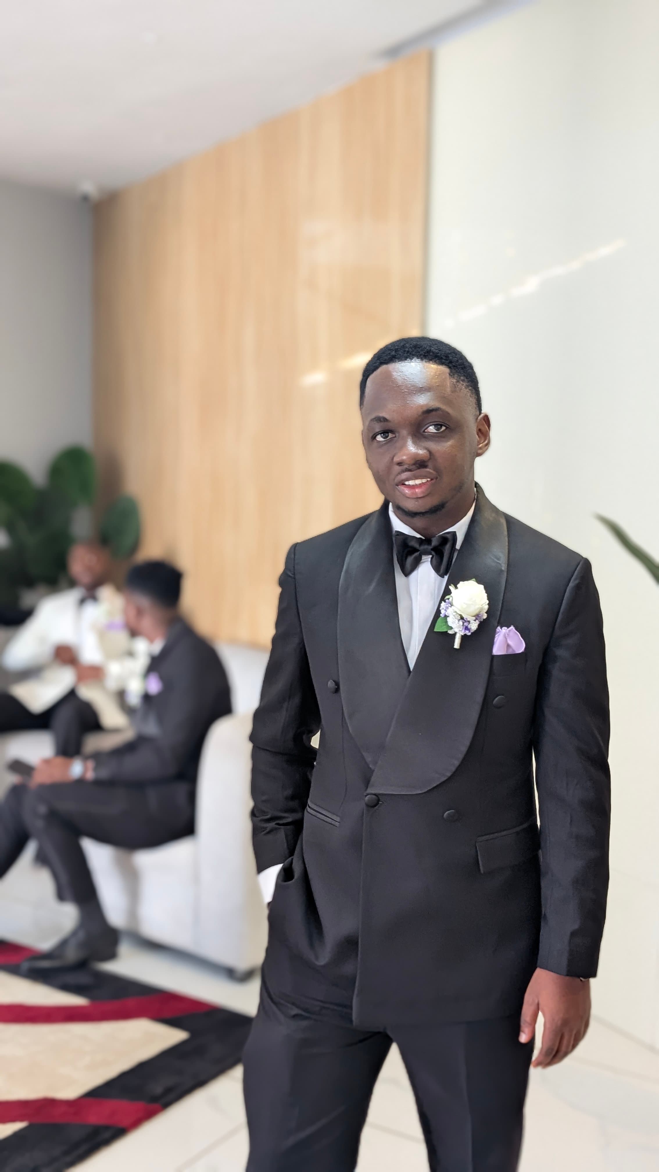 Man in a black tuxedo with a white boutonniere and purple pocket square posing indoors.