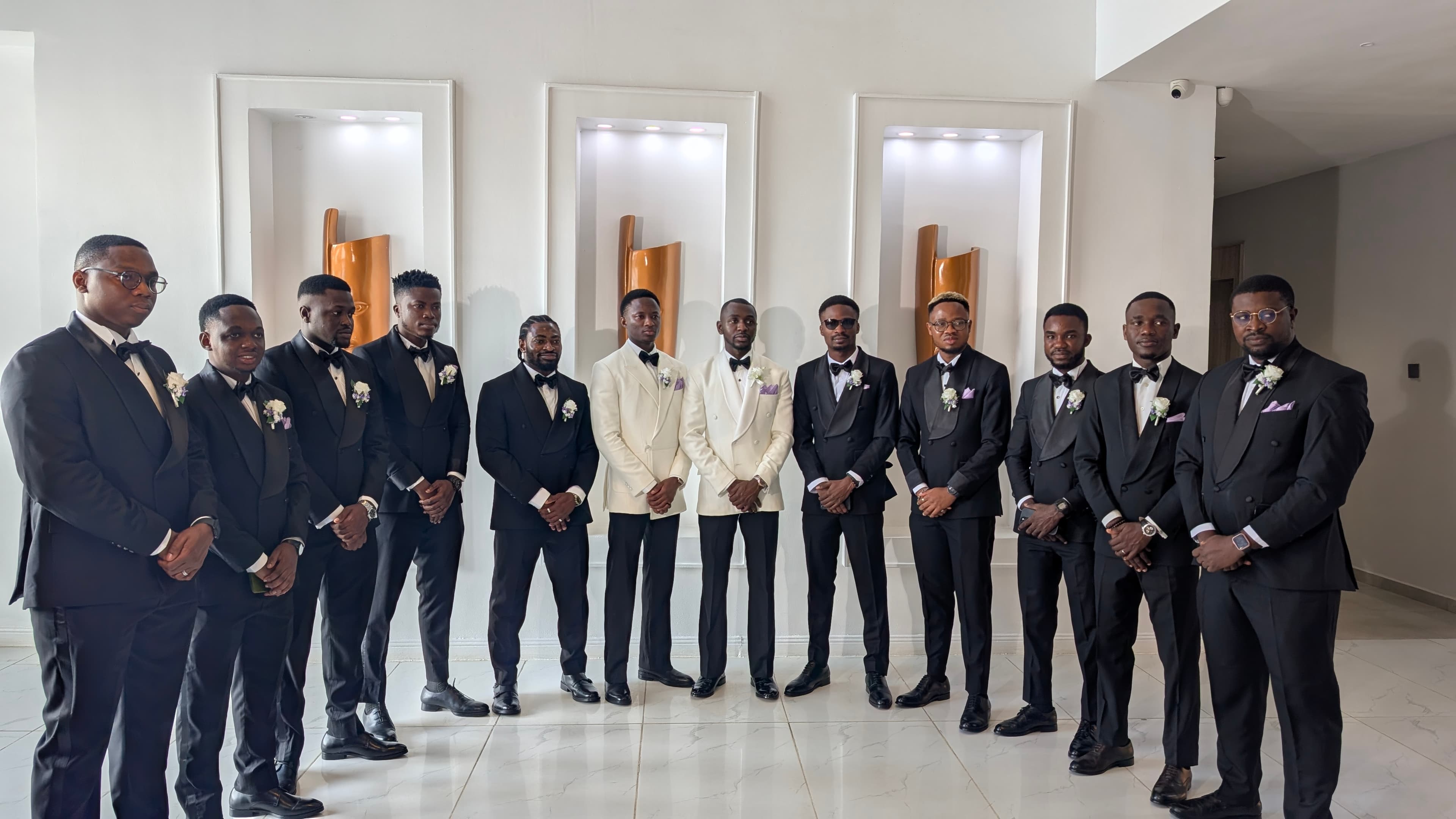 Twelve men in formal black and white tuxedos posing in a modern, brightly lit room.