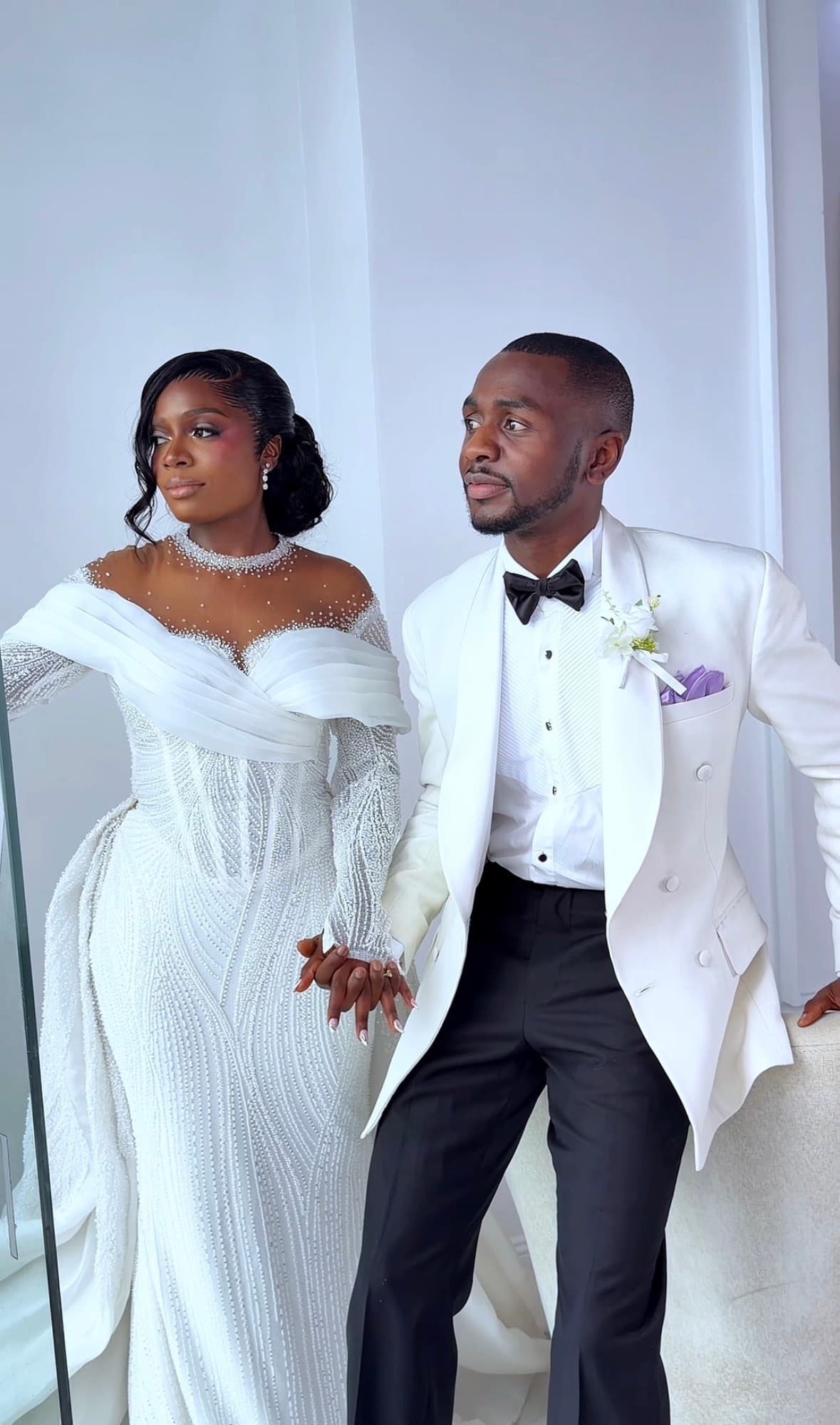 Black bride in beaded white gown and groom in white tuxedo holding hands, looking away.