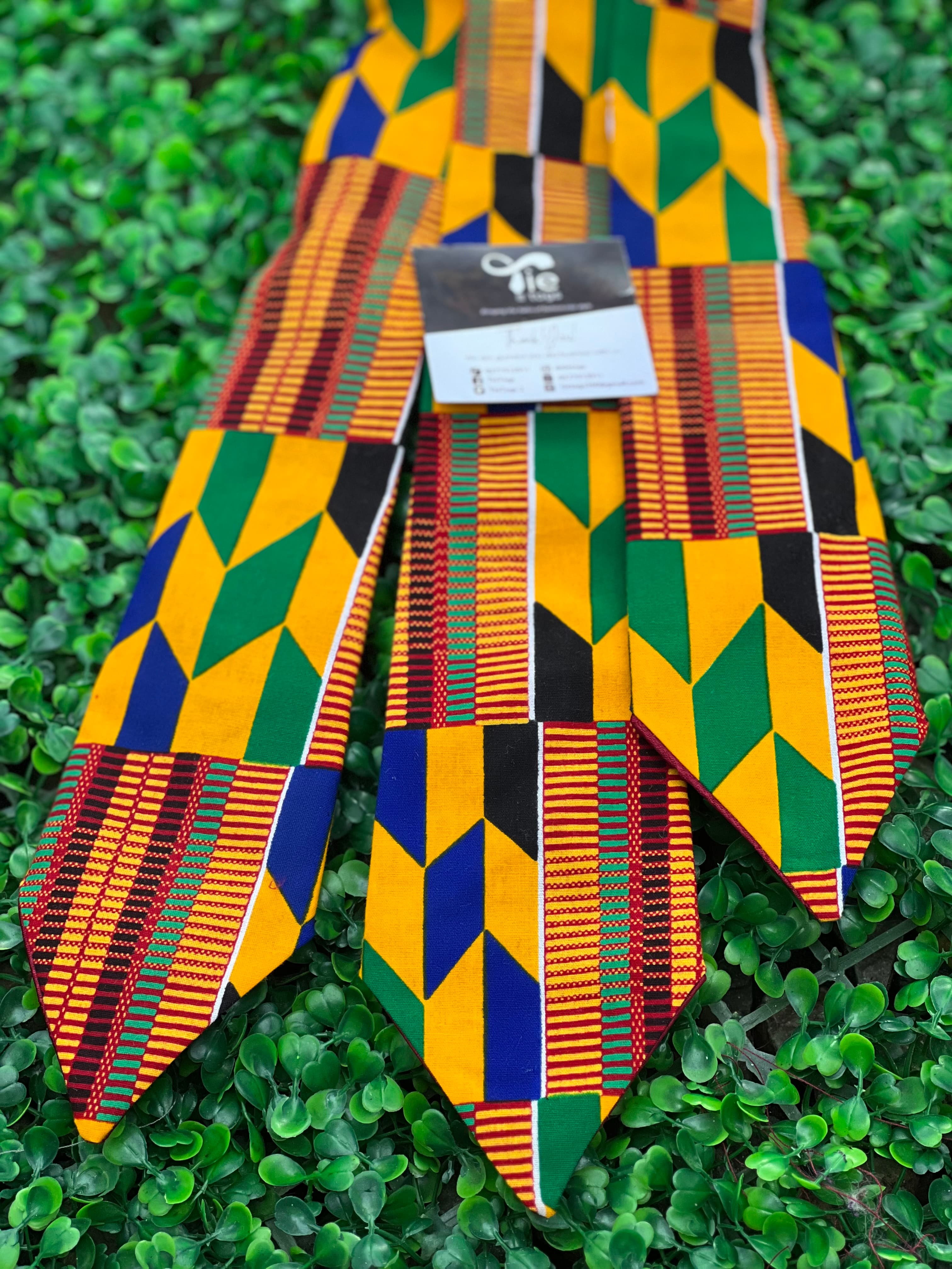 Three vibrant Kente cloth neckties with geometric patterns displayed on a green leafy background.