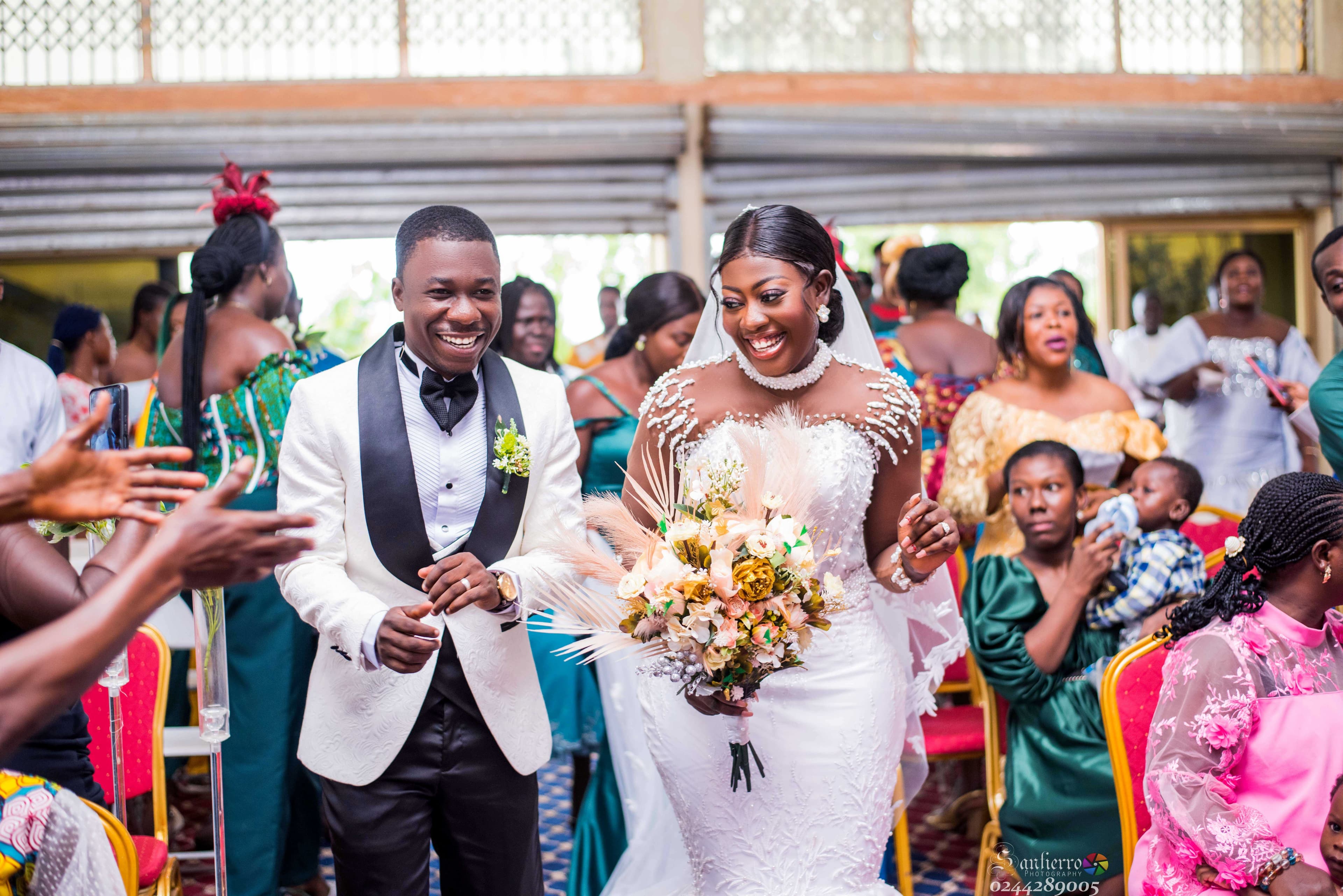 A joyful bride and groom walk down the aisle surrounded by cheering wedding guests.