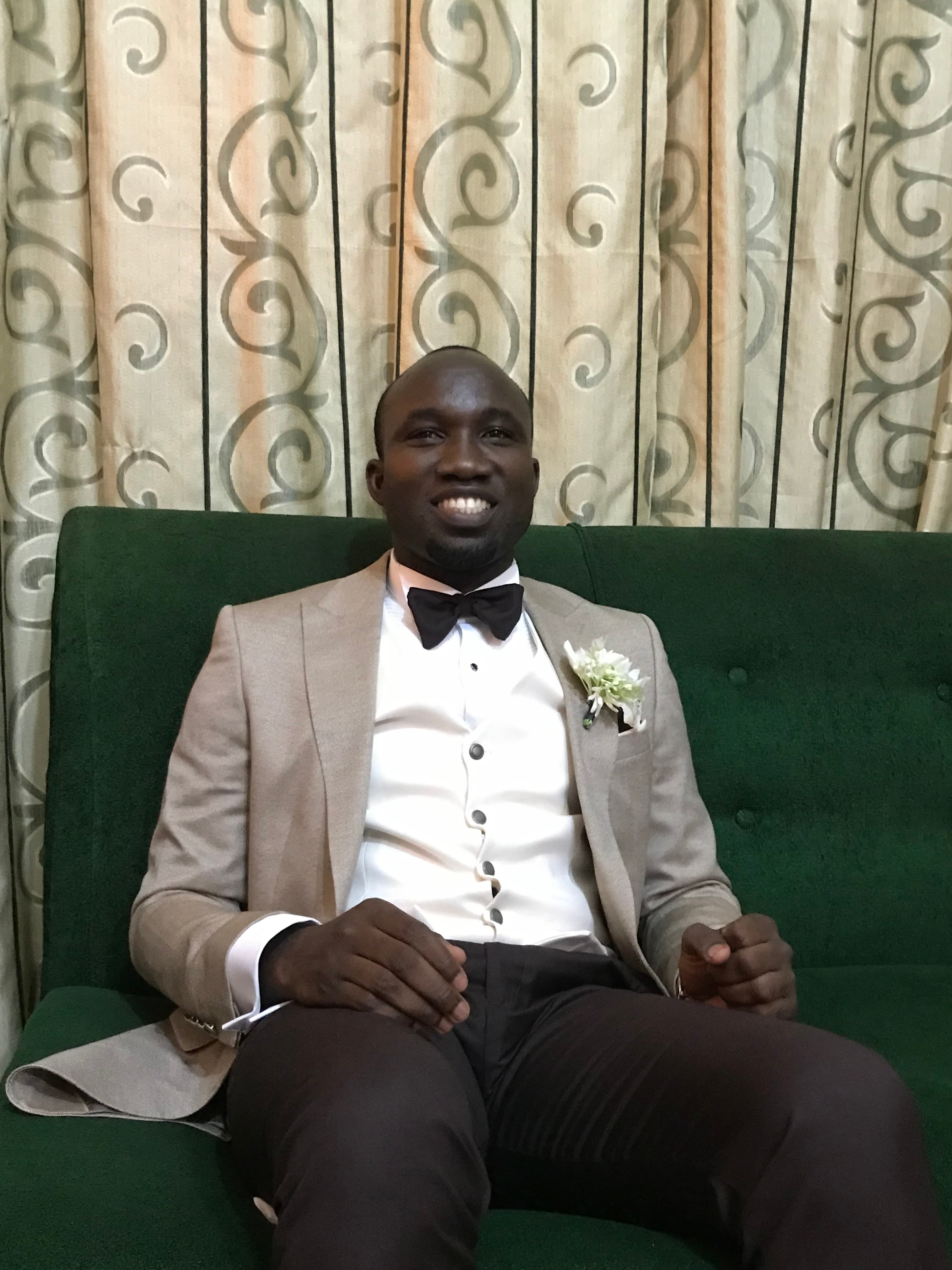 Smiling man wearing a tan blazer and black bow tie on a green couch.