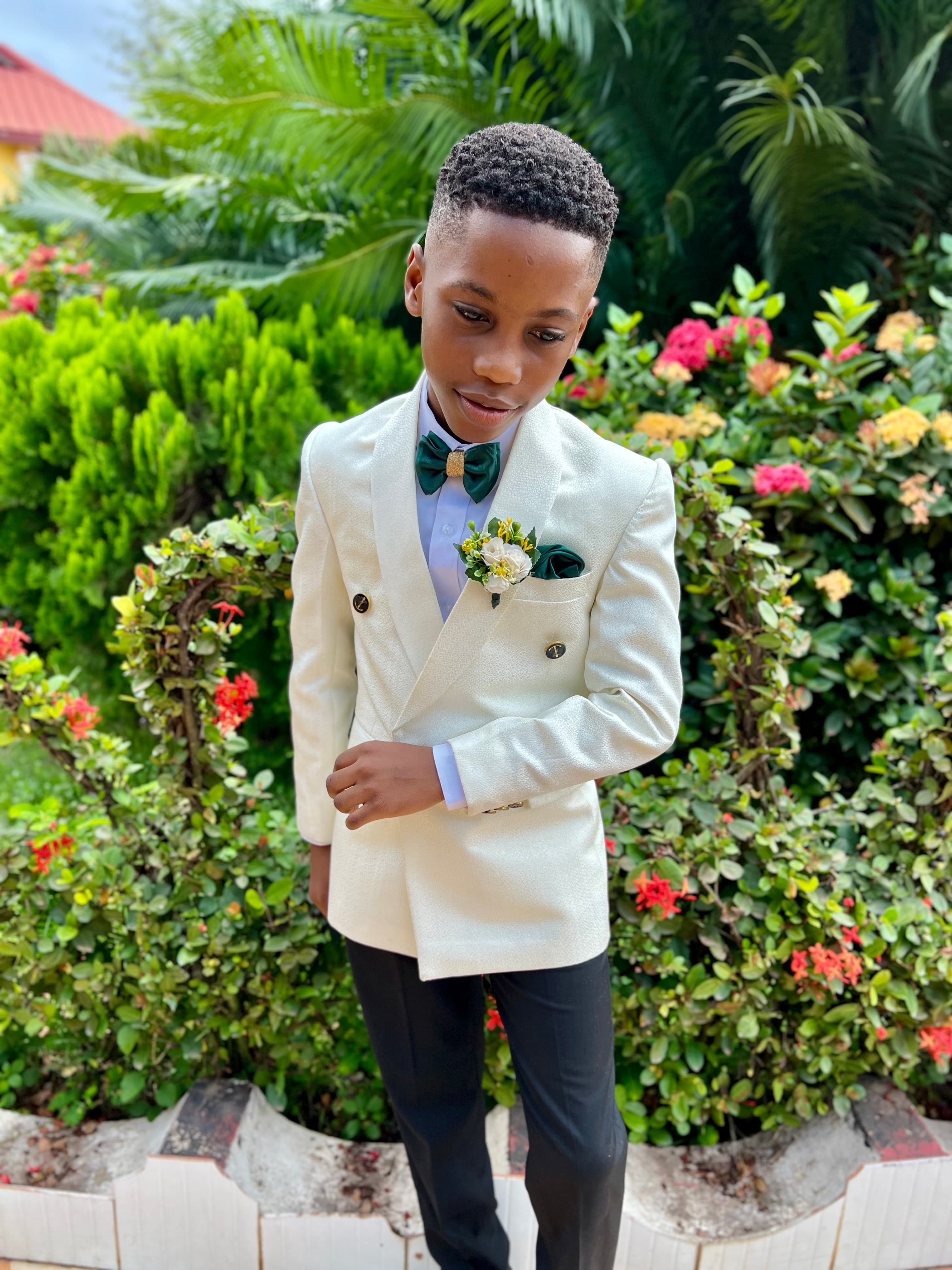 Young boy in a white double-breasted suit and green bow tie posing in a lush garden.