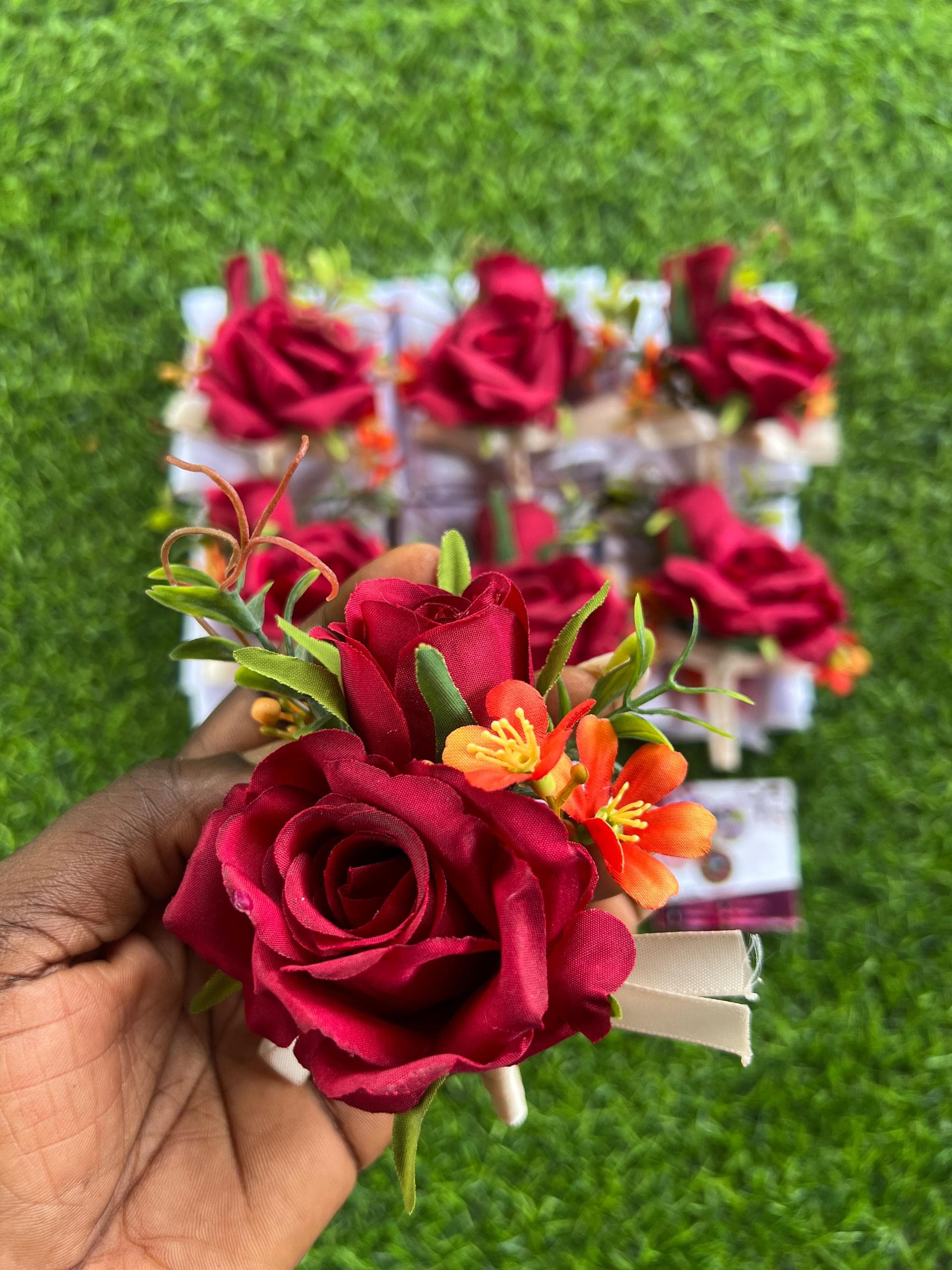 Hand holding a red rose boutonniere with orange flowers over a green grass background.