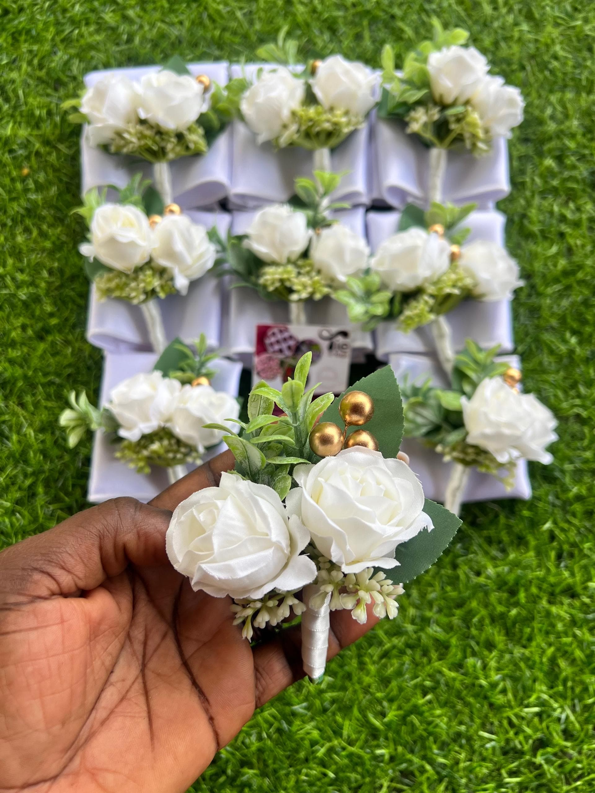 Hand holding a white rose boutonniere with gold beads over more boutonnieres on green grass.