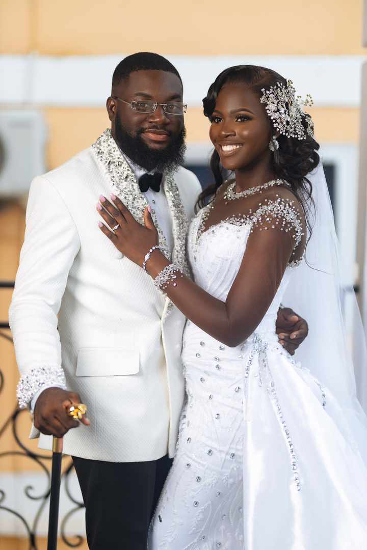 Smiling Black bride and groom in ornate white wedding attire posing for a formal portrait.