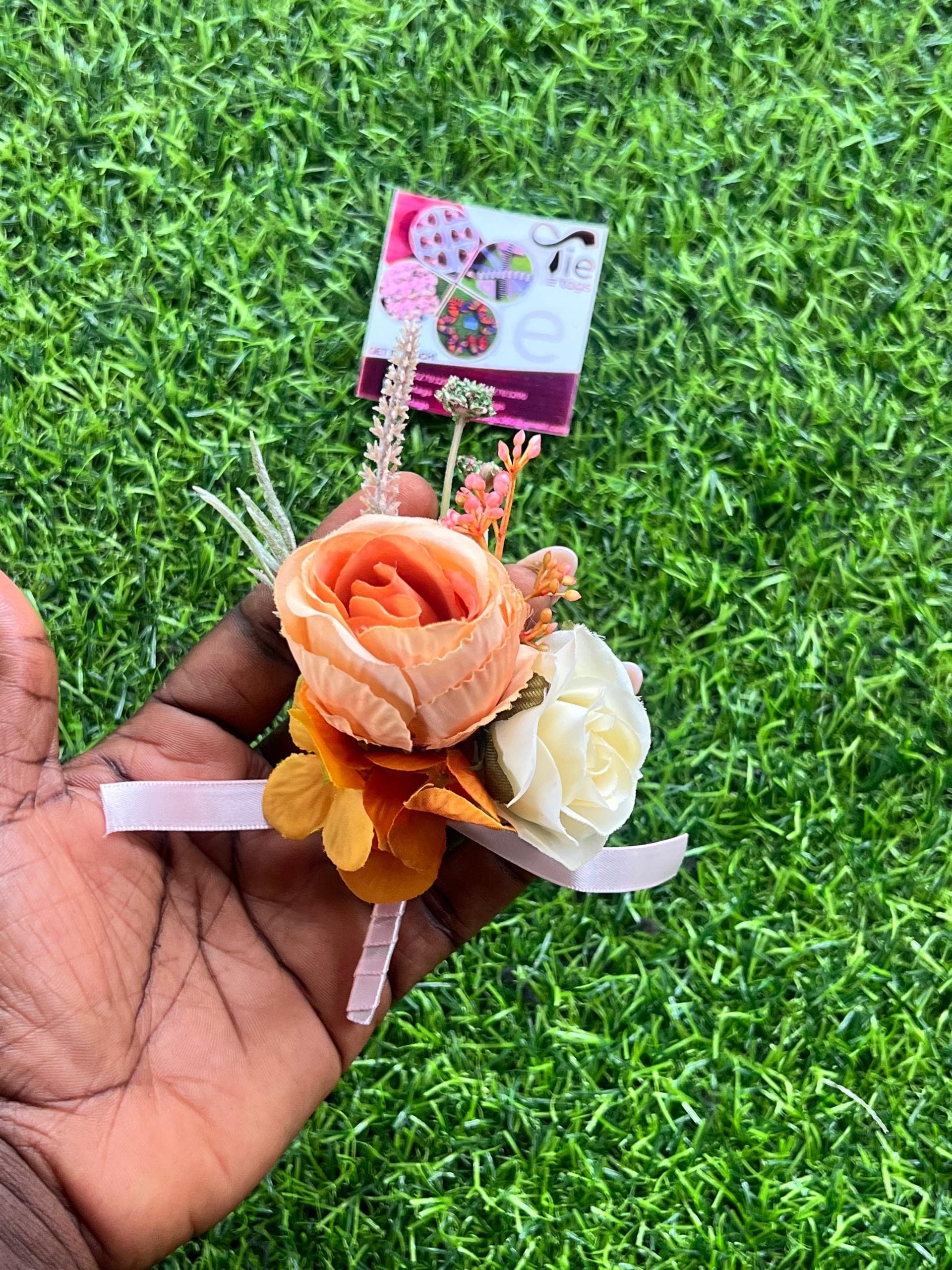 Hand holding a peach and white rose boutonniere with a pink ribbon over green grass.